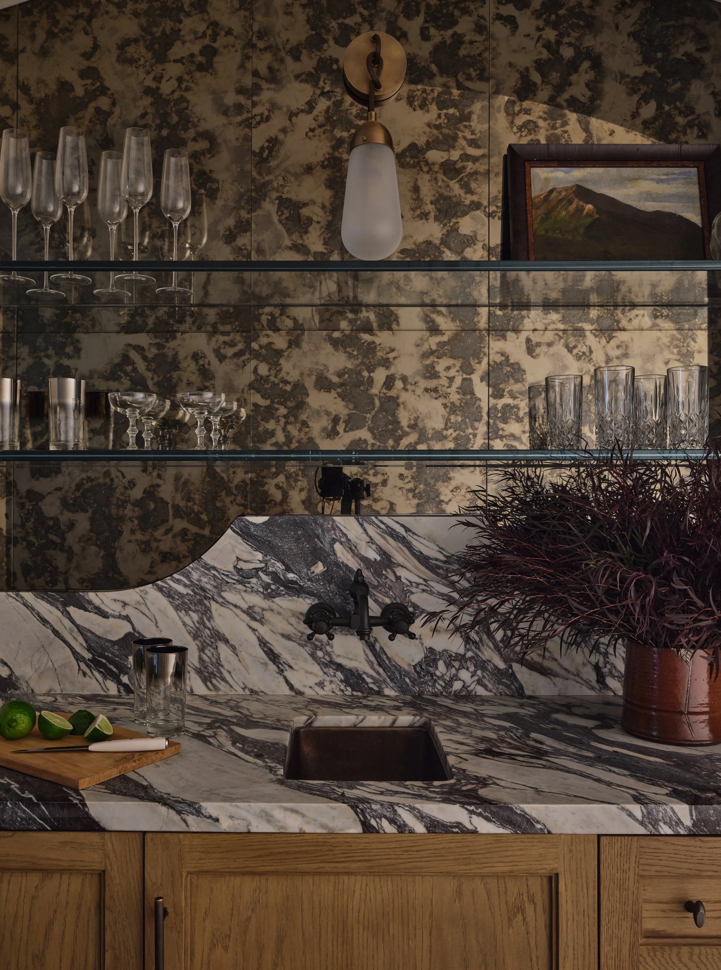A kitchen with a marble countertop and backsplash, a black faucet, top shelves with glassware, a wooden cabinet, a potted plant, and a wall-mounted light fixture