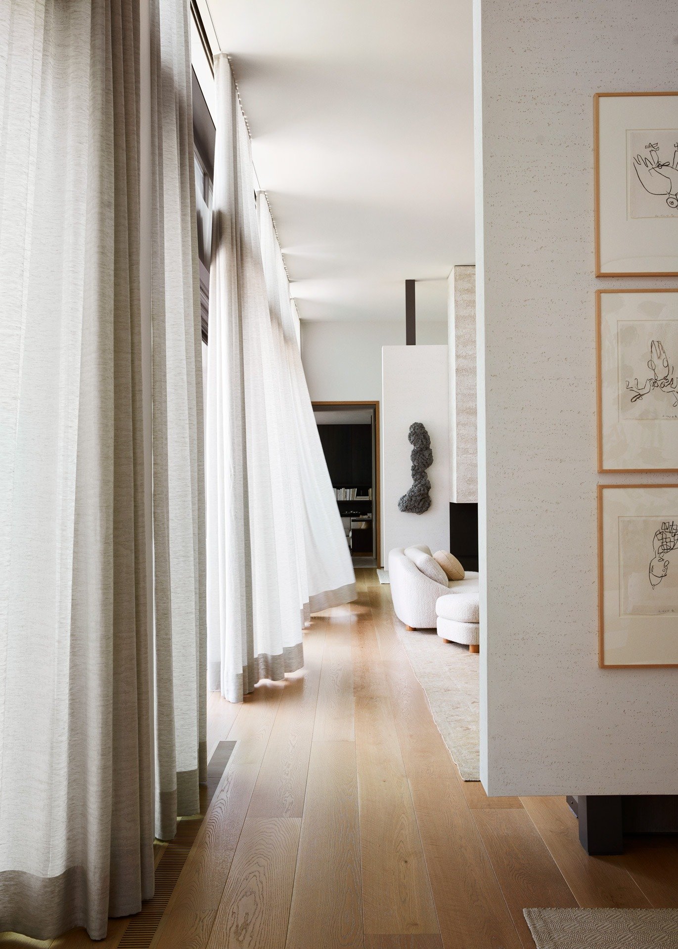 Bright living room with large window covered by off-white curtains, light-colored wood flooring, a white curved sofa, and artwork on the walls.