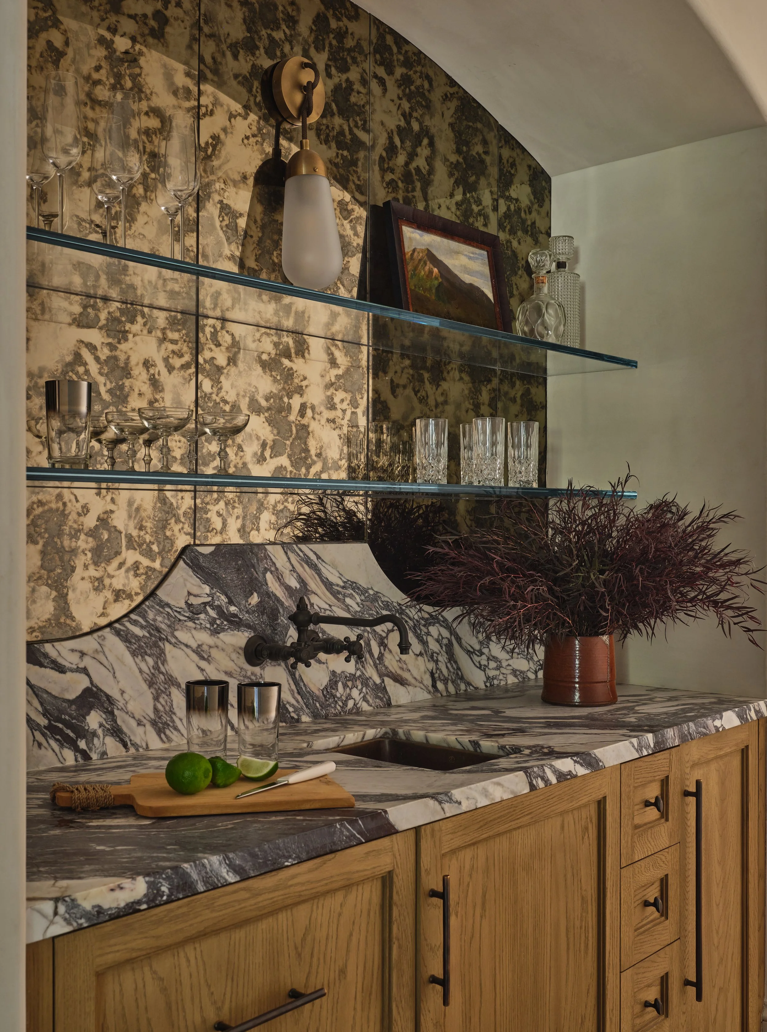 Kitchen with a marble countertop and backsplash, wooden cabinets, glass shelves with glasses and artwork, and a potted plant with dark purple leaves.
