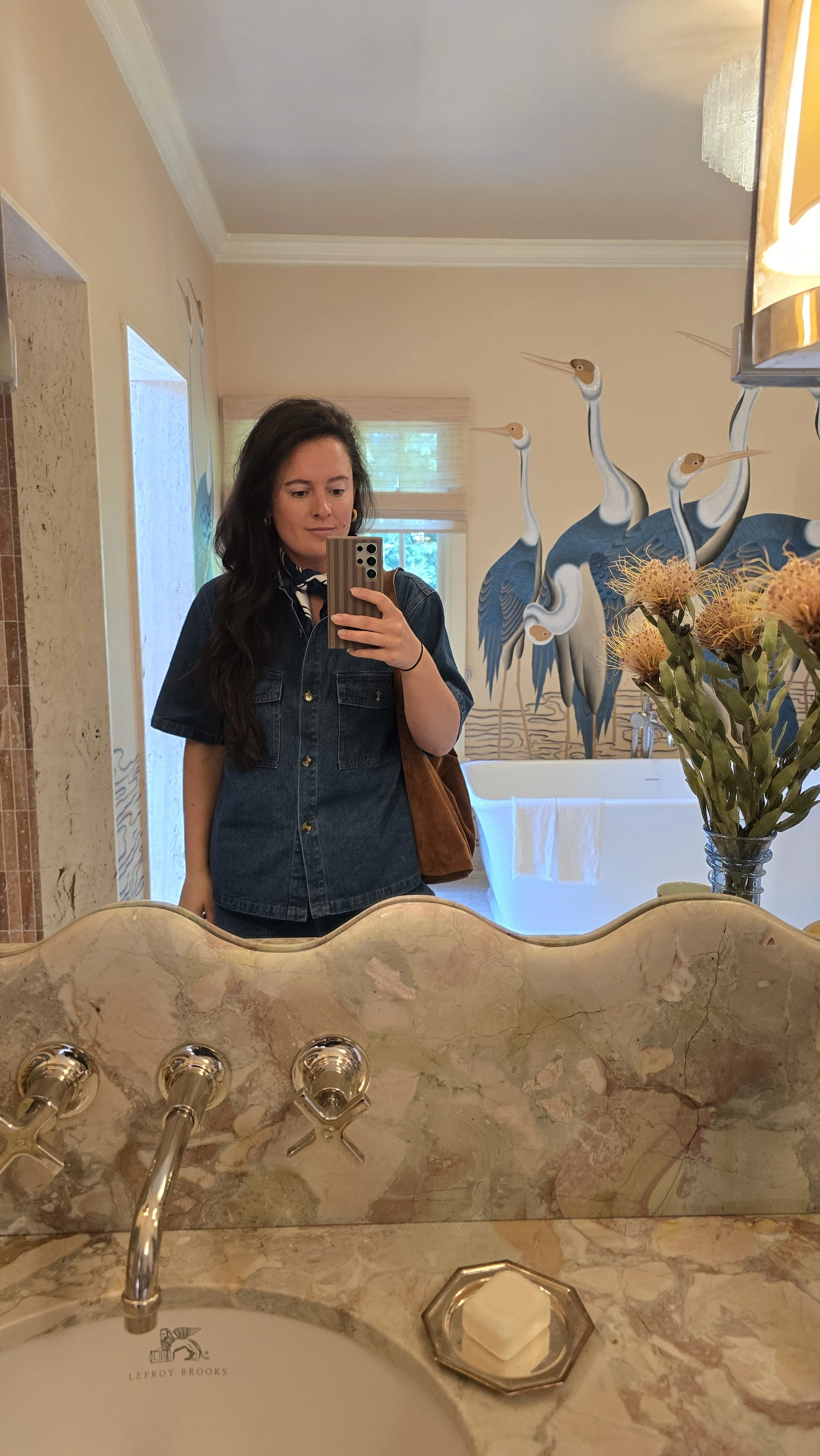 Interior Deisgner Samantha Riccio  taking a mirror selfie in a bathroom with a mural of cranes, flowers in a vase, and a countertop with soap.