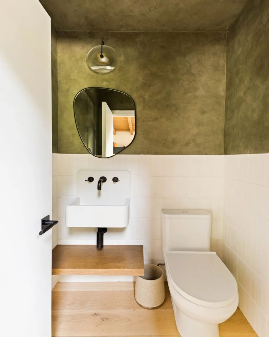 A powder room with a stacked white cement tile wainscot and olive green plaster walls.  There is a white concrete sink with a floating wood shelf underneath.  An organic shaped mirror and a allied maker sconce is above the sink.