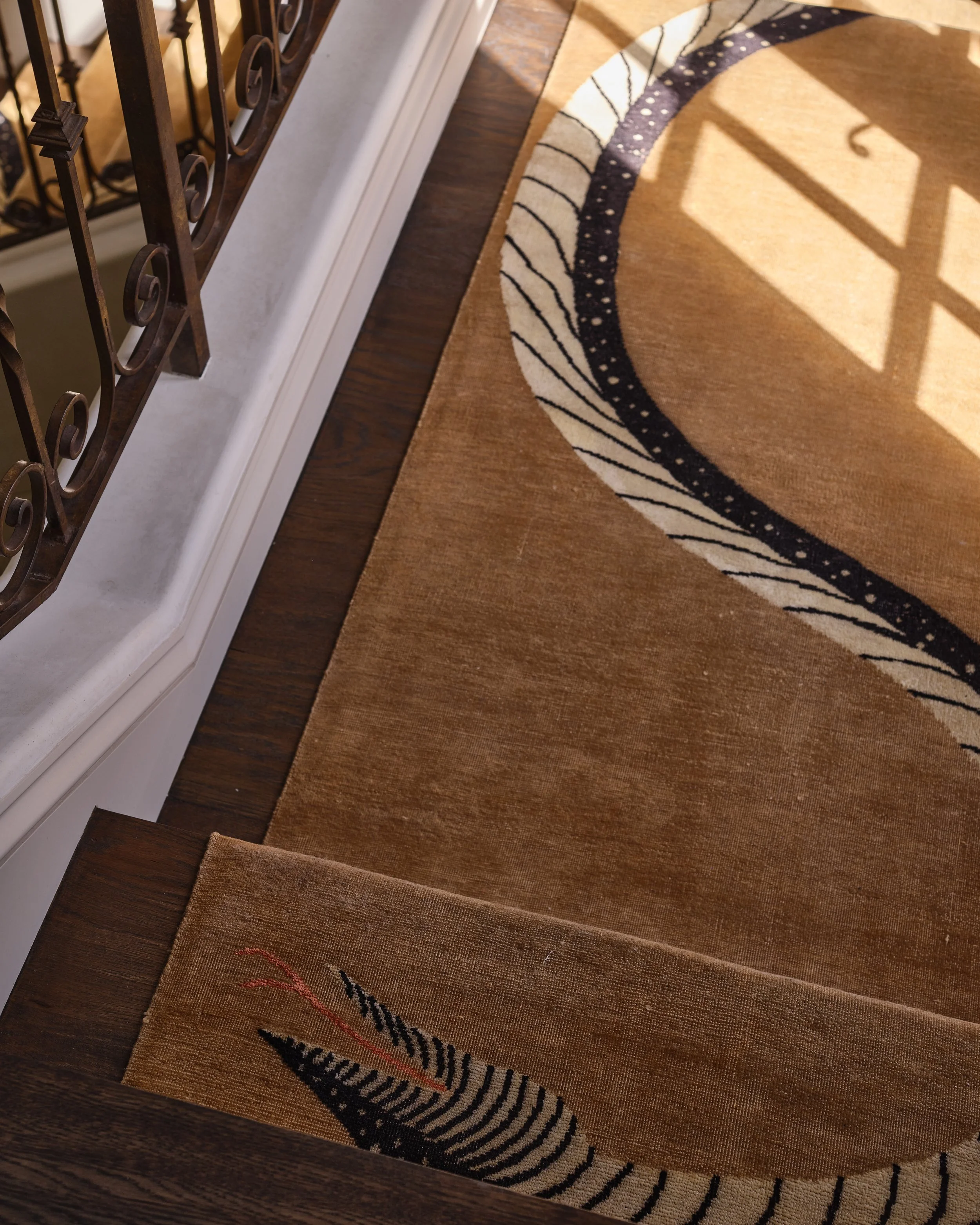 A brown and beige patterned area rug with black and white curved lines, on a wooden floor near a staircase with a metal railing.