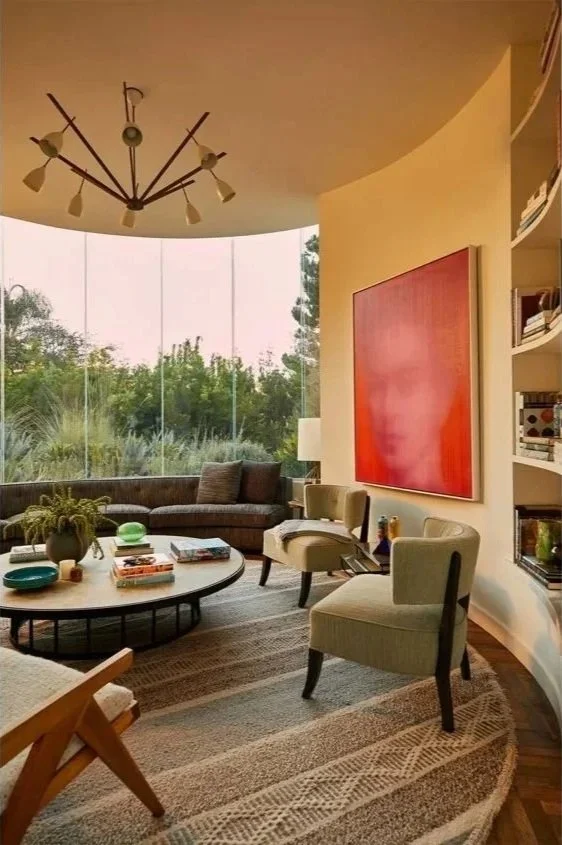 Living room with large window view of greenery, a dark curved sofa, four beige chairs, a round coffee table with books and decorative items, a large red painting on the wall, a modern chandelier, and a curved white built-in bookshelf.