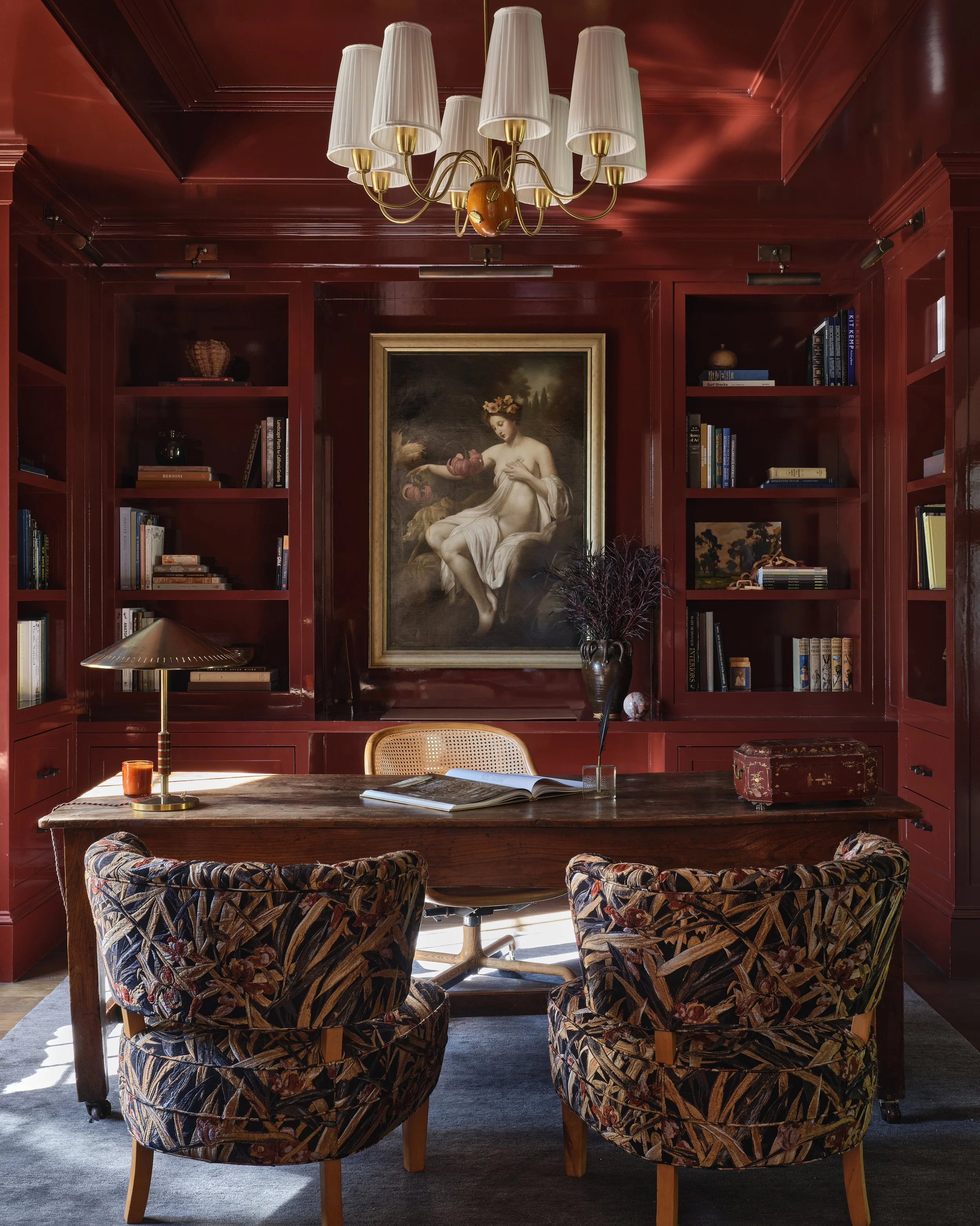 A study room with red wood-paneled walls, a wooden desk, two patterned armchairs, a bookshelf, a large framed classical painting of a woman, a desk lamp, a vase with dark branches, and a chandelier with white lampshades.
