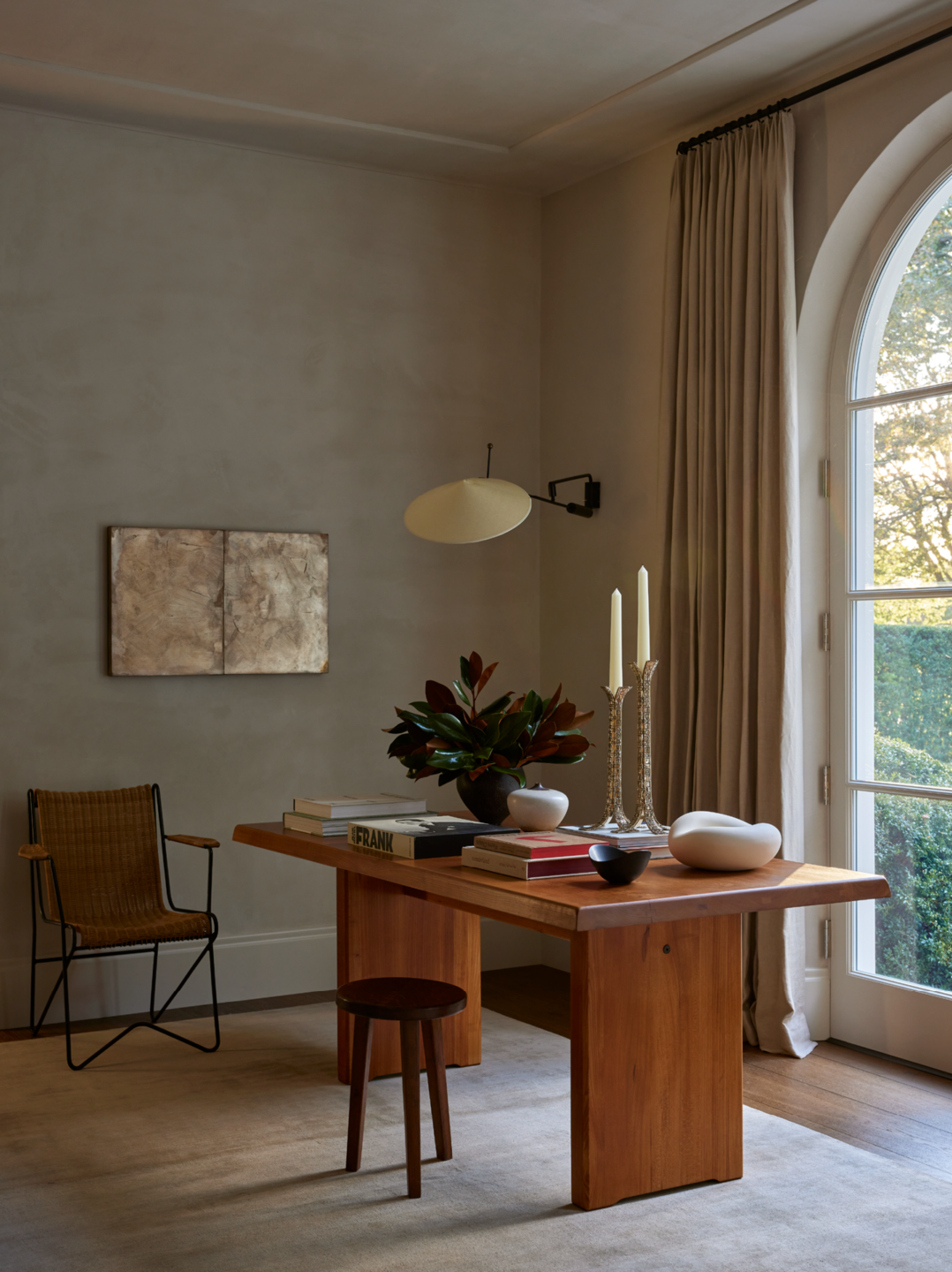 A cozy dining or work area with a wooden table, decorative vases and candles, a small stool, a woven chair, a wall-mounted lamp, and an arched window with beige curtains allowing natural light into the room