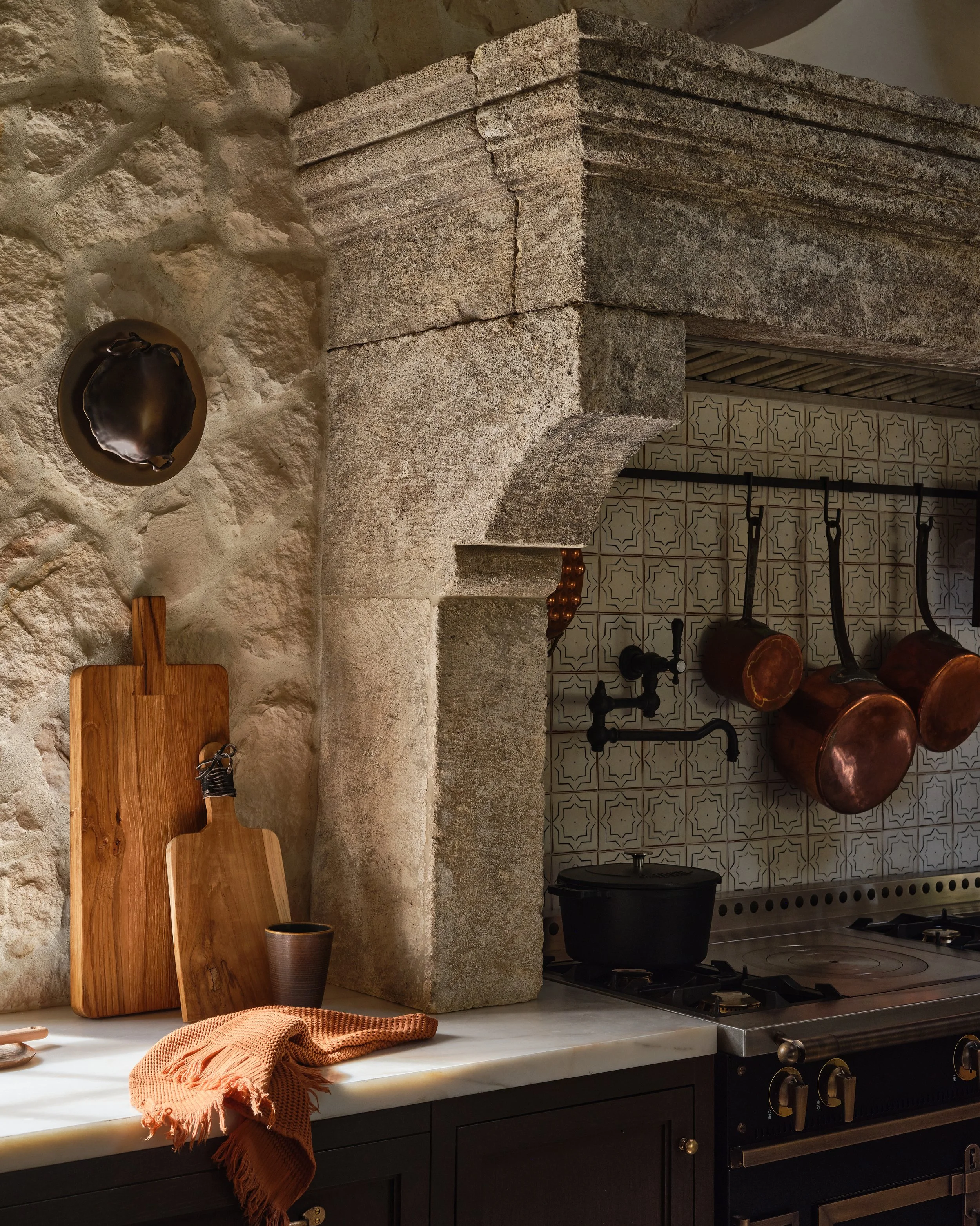 Rustic kitchen with stone and tiled walls, copper pots hanging, and a stovetop with black cabinetry. Wooden cutting boards, a ceramic cup, and an orange cloth on the marble countertop.