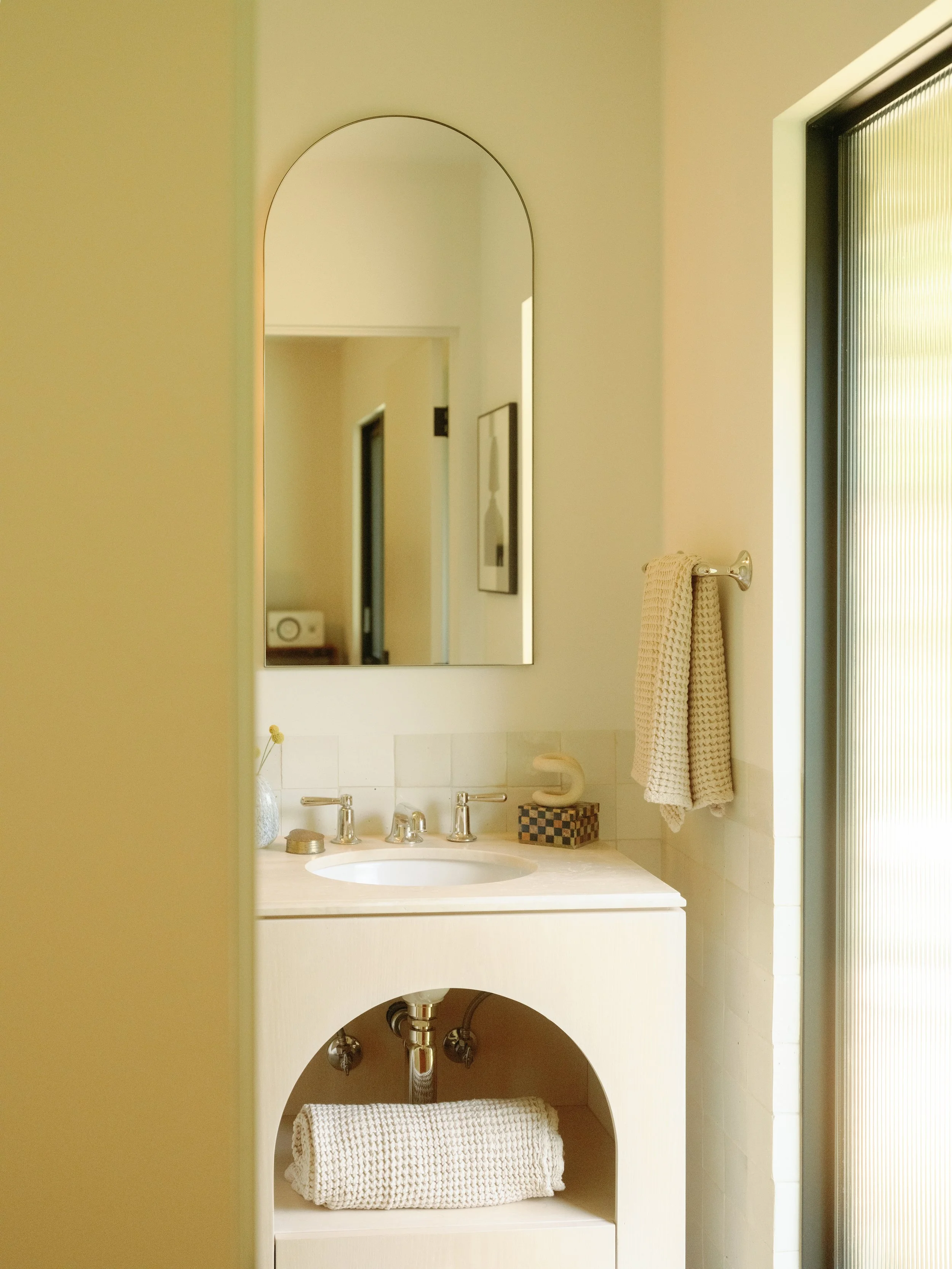 A bathroom vanity with a white countertop, a round sink, and a three-hole faucet. There is a decorative sculpture and a checkered box on the counter, with a towel ring and towel on the wall. A large window or door with vertical ridges is to the right. A mirror hangs above the vanity, reflecting an interior hallway.