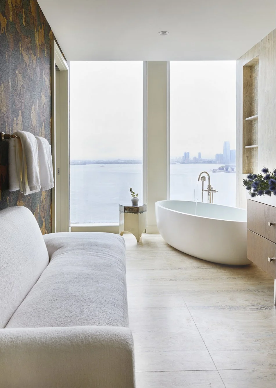 Modern bathroom with large windows overlooking a city skyline and water, featuring a freestanding bathtub, a small side table with a plant, a blanket on a couch, and towels hanging on the wall.