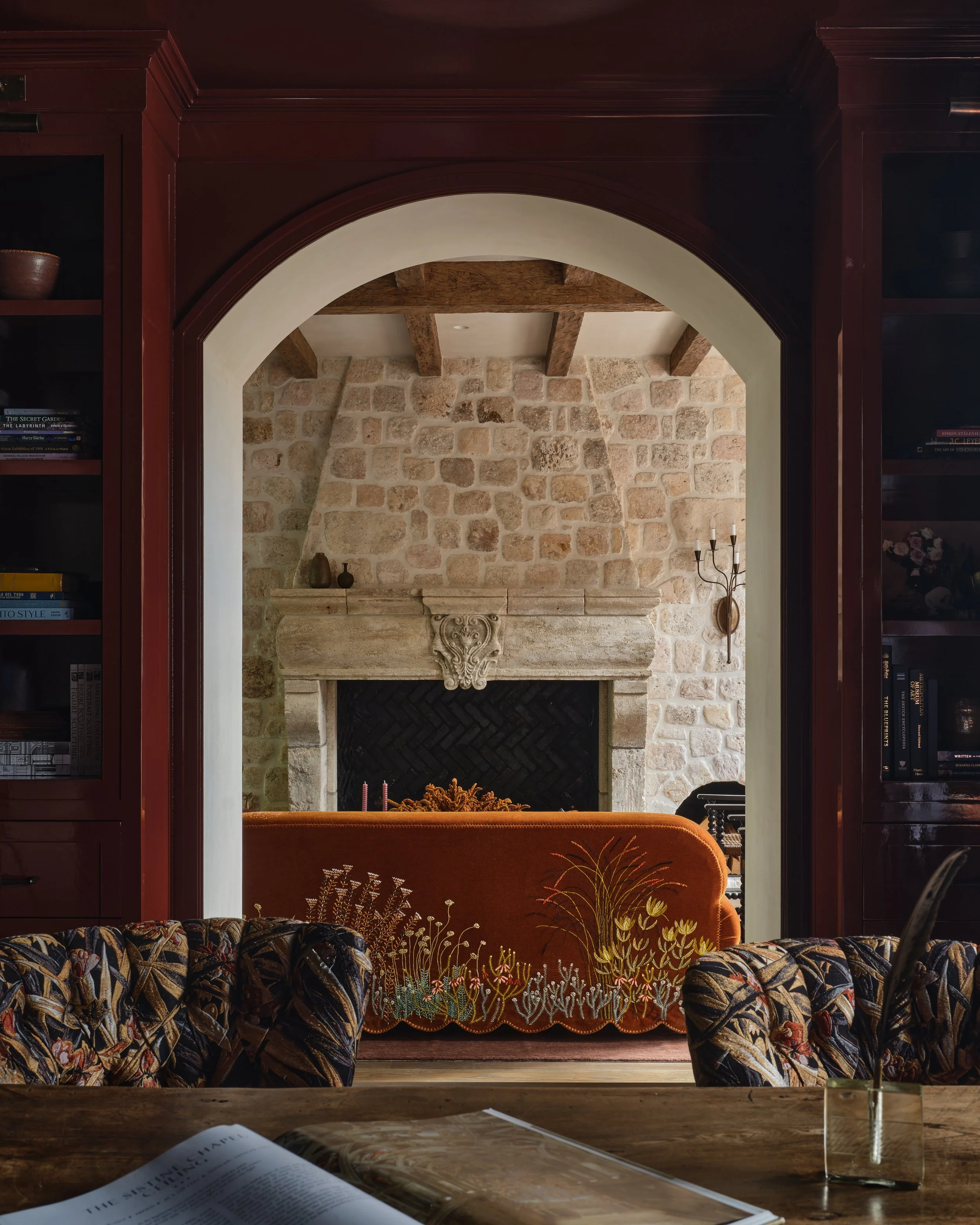 Interior view of a room with a stone fireplace, wooden beams, a vintage orange velvet sofa with floral embroidery, and a book on a wooden table in the foreground.