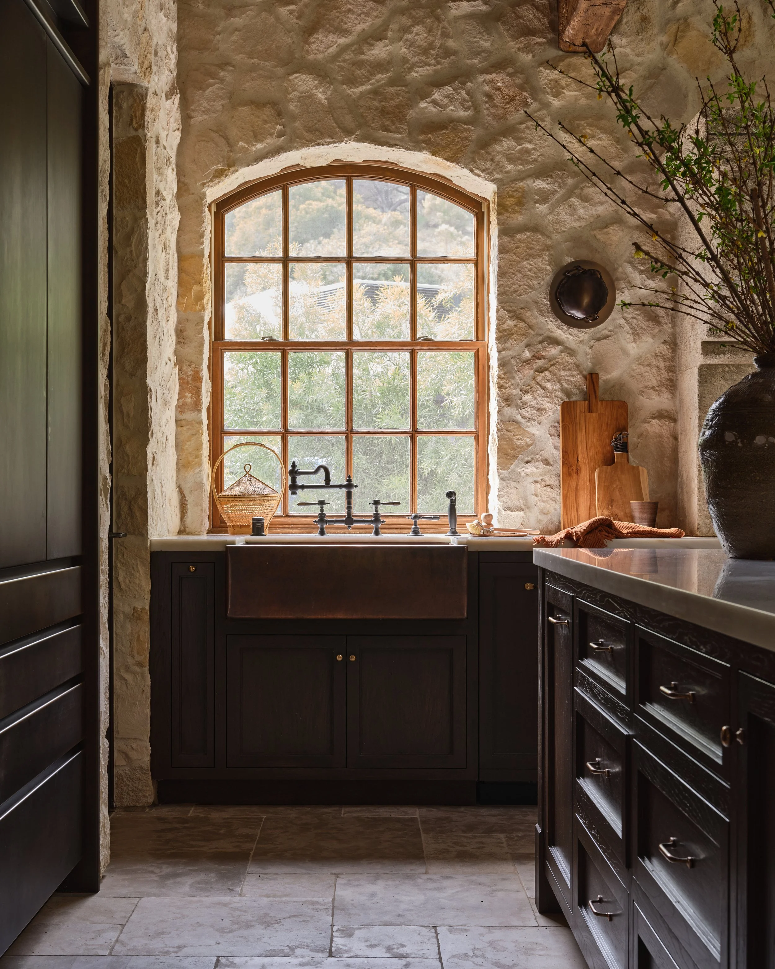 A rustic kitchen with stone walls, a large window with wooden frames overlooking greenery, a farmhouse sink with a black finish, wooden cutting boards, and a large vase with branches.