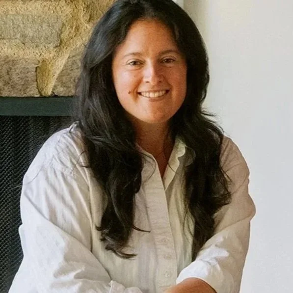 A woman with long black hair and a white button-up shirt, smiling, sitting indoors near a fireplace.