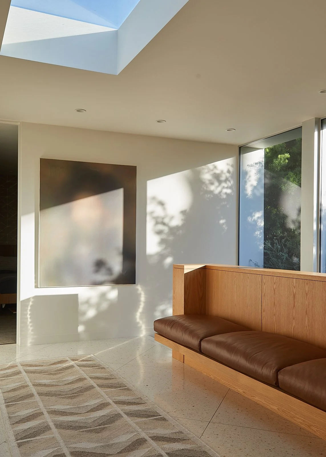 Modern interior with a wooden bench with brown leather cushion, a large window with outdoor greenery, a framed artwork on the wall, a ceiling skylight, and natural sunlight casting shadows.