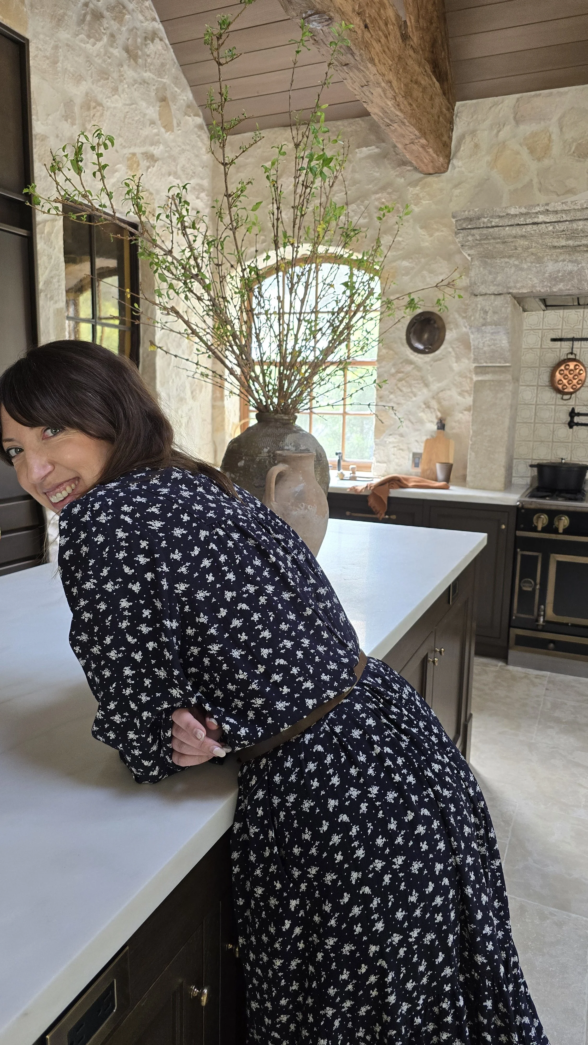 Interior Designer Ariane Vidaurre leaning on a kitchen counter in a rustic kitchen with stone walls, large vase with branches, and a window with natural light.