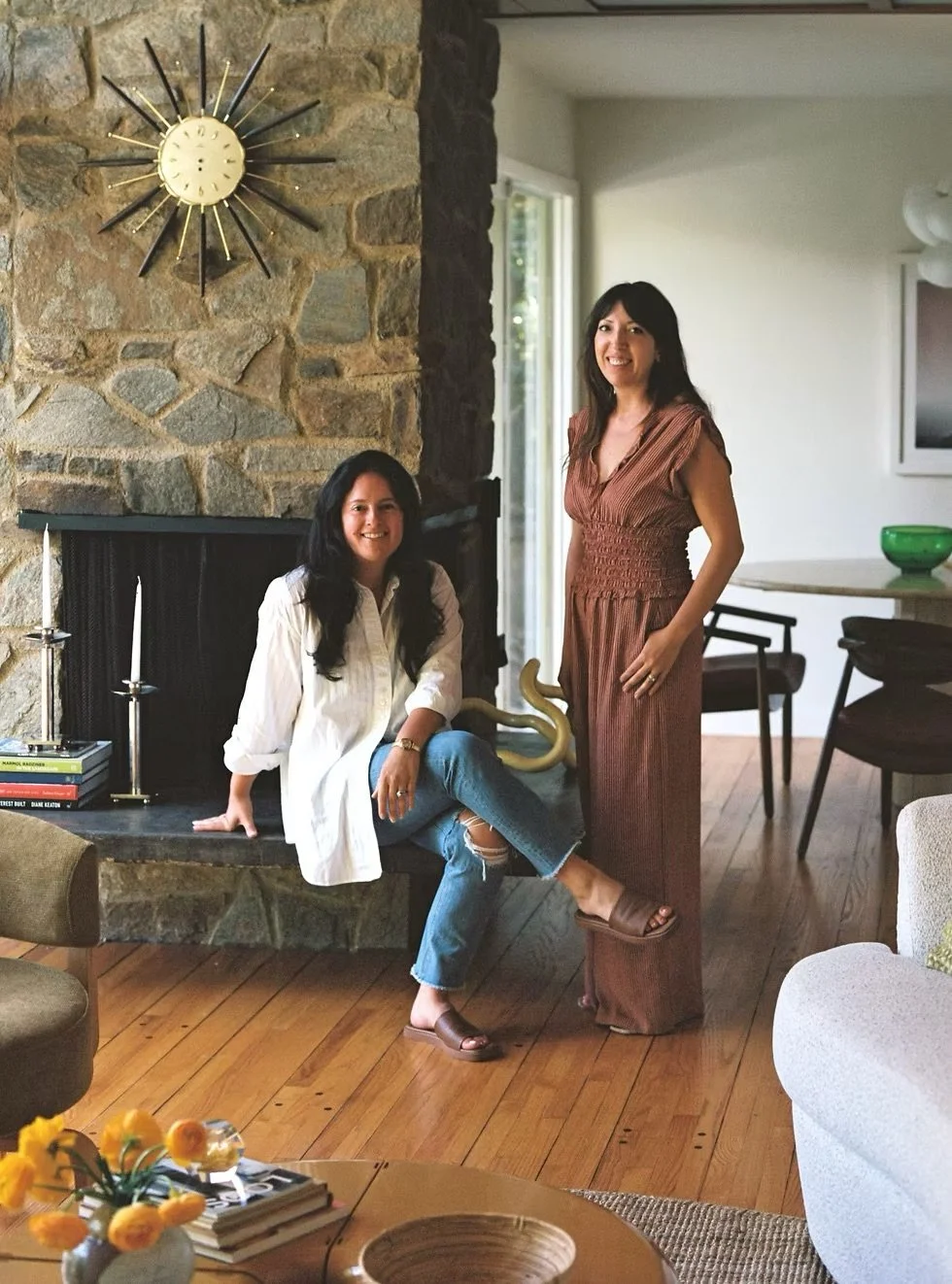 Two women in a living room with a stone fireplace. One woman sits on the fireplace ledge, and the other stands next to her. The room has wooden floors, a TV, and a window letting in natural light.