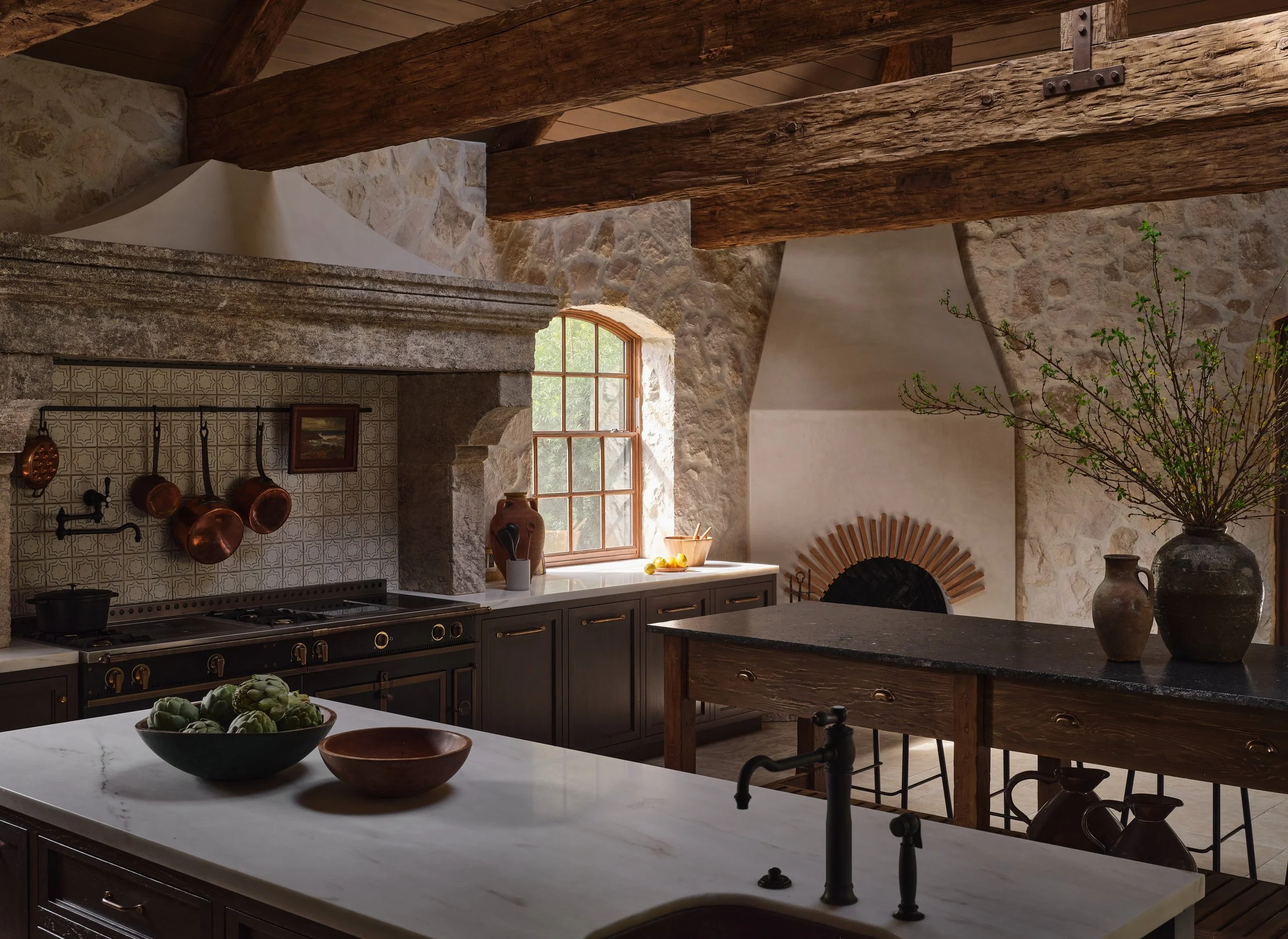 Rustic French country style kitchen with stone walls, wooden ceiling and double islands.  A black la cornue range is topped with a giant antique stone hood, copper pots and a small antique painting hang above.