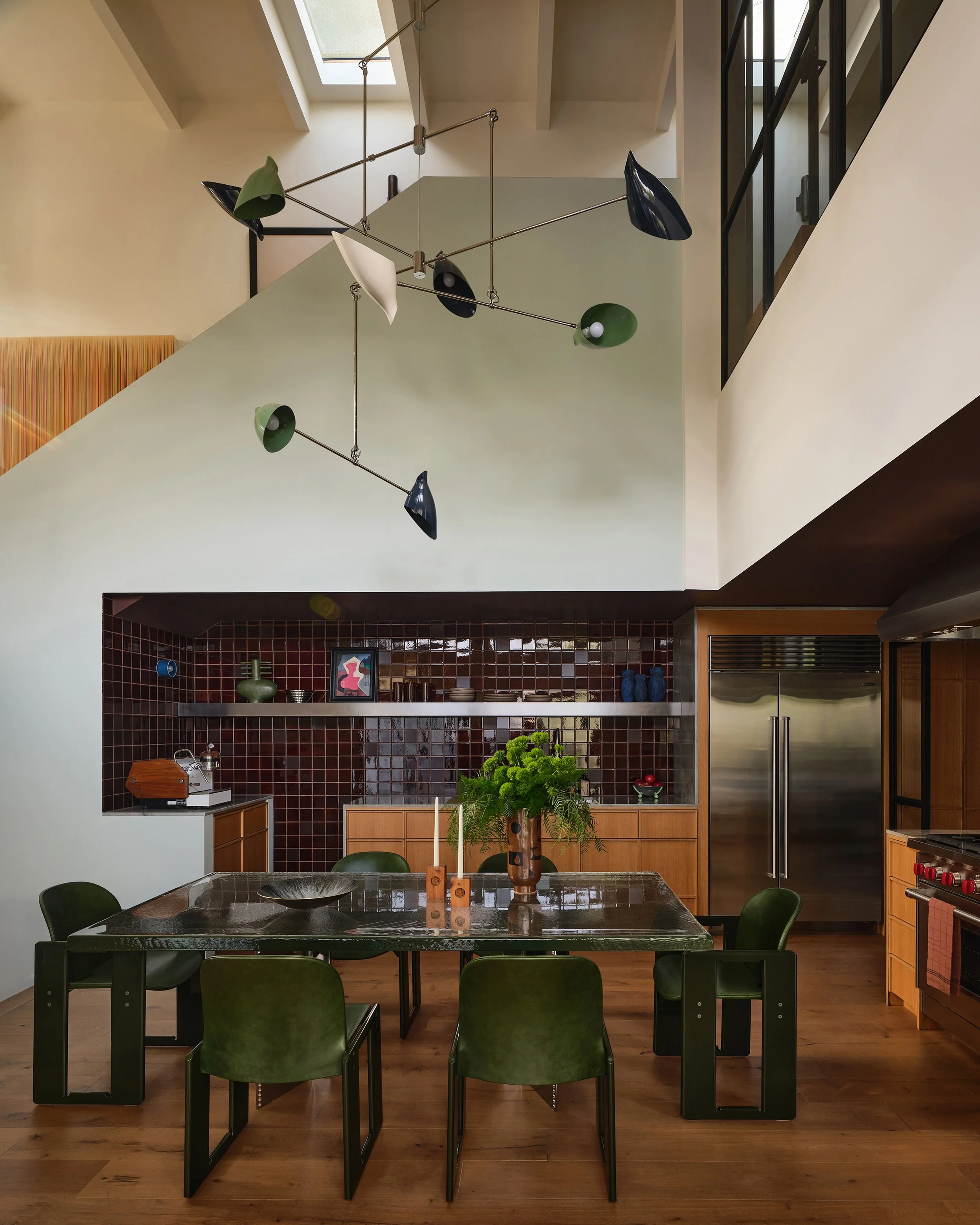 Comedian Anthony Jeselnik’s home by Riccio Vidaurre Design – A kitchen vaulted ceilings, a cast glass dining table and green dialogo dining chairs by Tobia Scarpa with a David weeks chandelier above. Brown ceramic tile backsplash and oak cabinetry.