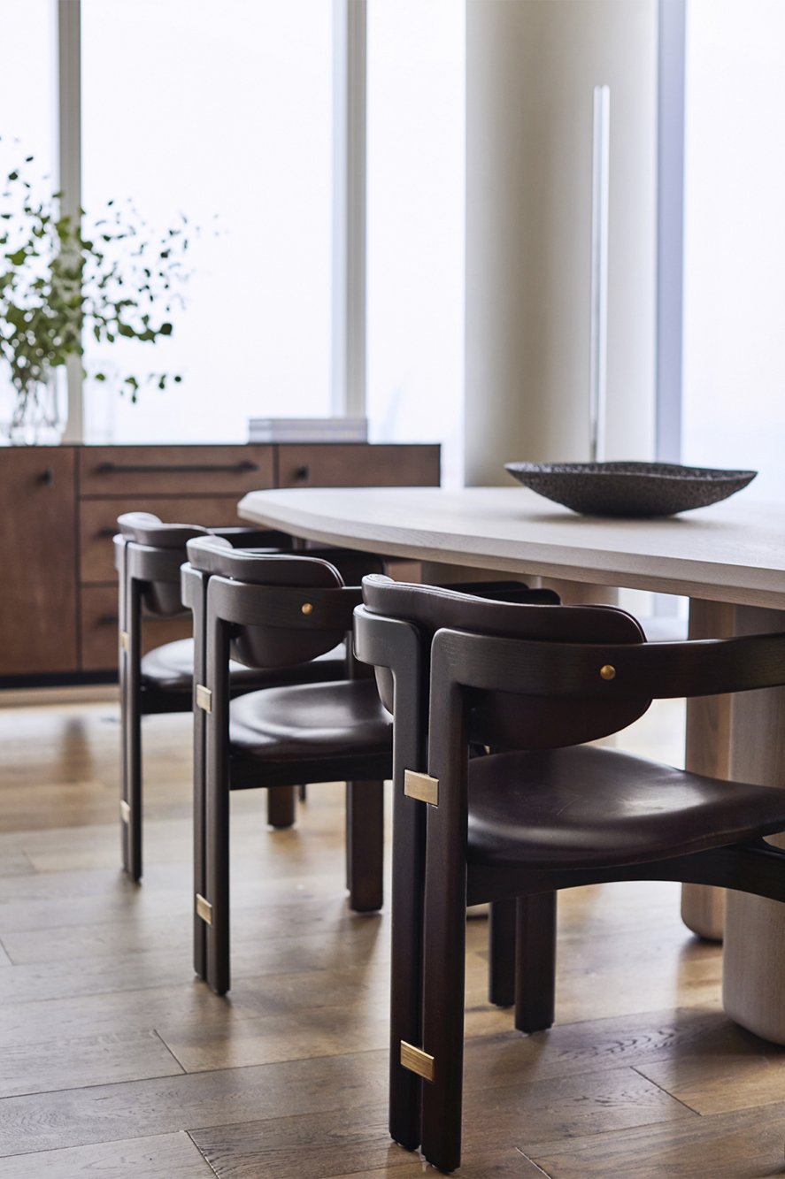 Modern kitchen with a light-colored dining table and three dark wood chairs with brass accents, a bowl on the table, and a wooden sideboard with a plant and books in the background, large windows providing natural light.