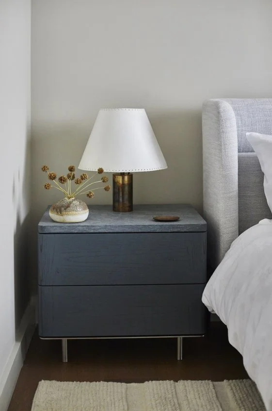 A bedroom nightstand with a lamp, decorative vase with dried flowers, and a small tray, next to a bed with a gray upholstered headboard.