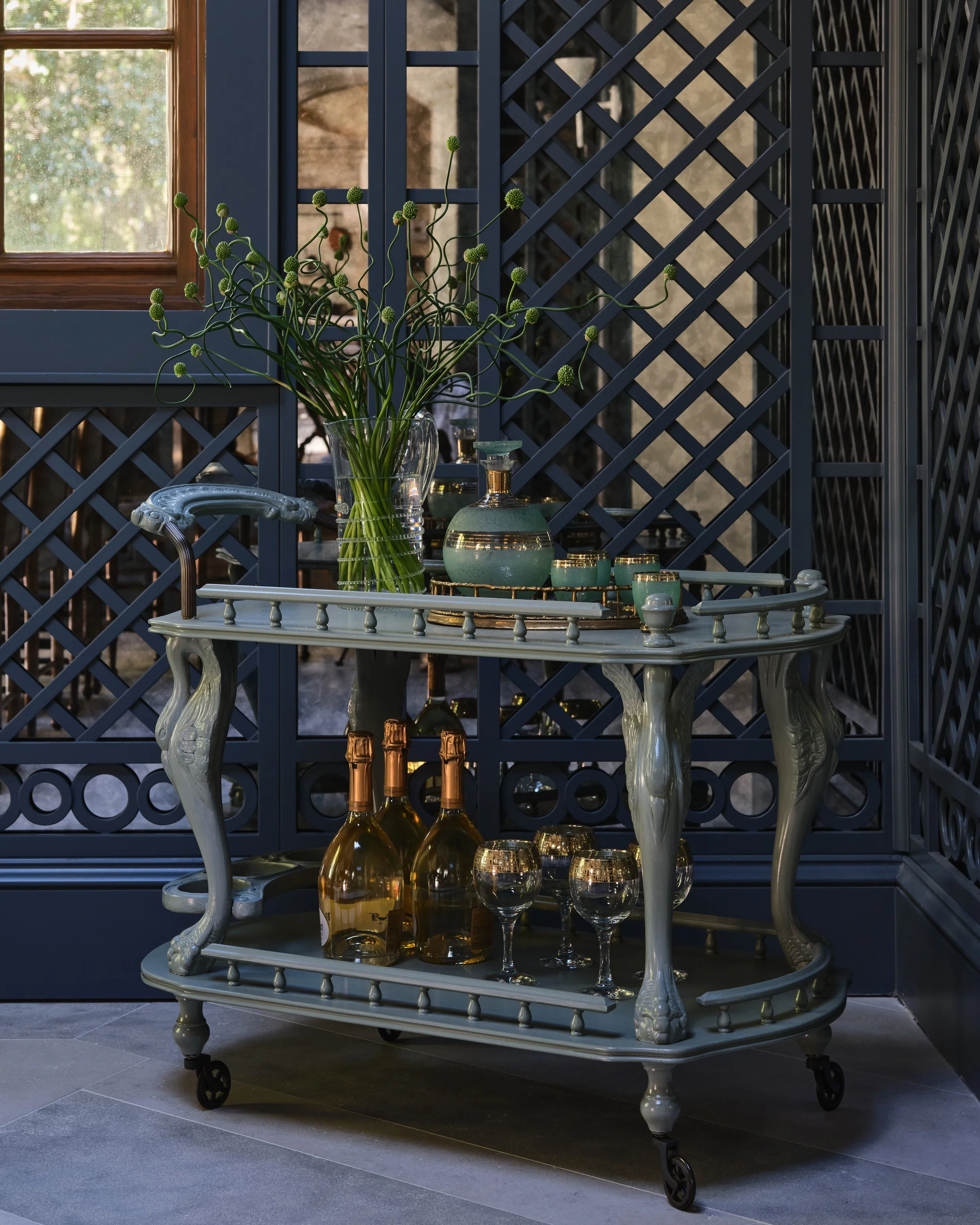 A vintage-style serving cart with a light green finish, ornate curved legs, and wheels, positioned on a tiled floor. On the top shelf, there is a large glass vase filled with tall, curly green plants with round buds and a matching green and gold ceramic decanter set. The bottom shelf holds three bottles of champagne and four wine glasses with gold rims. In the background, there is a window with wooden framing and a decorative dark blue lattice wall.