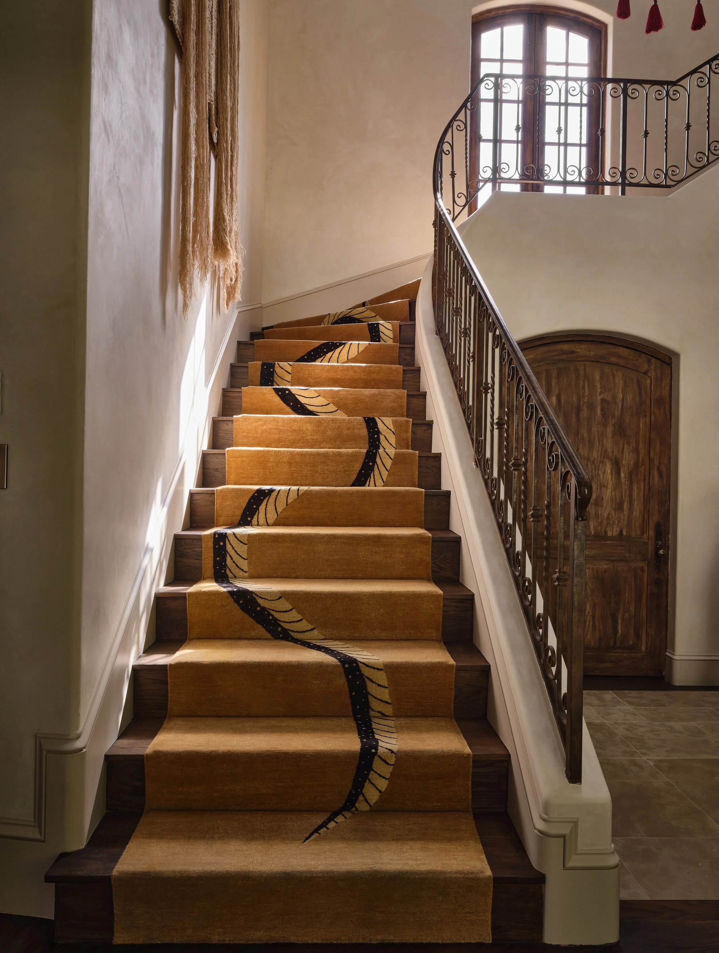 Interior view of a staircase with a beige and black patterned runner, wooden steps, and ornate black metal railing. A large arched window with grid panes lets in natural light. A wooden door is at the base of the stairs, and beige walls and ceiling with red tassels are visible.