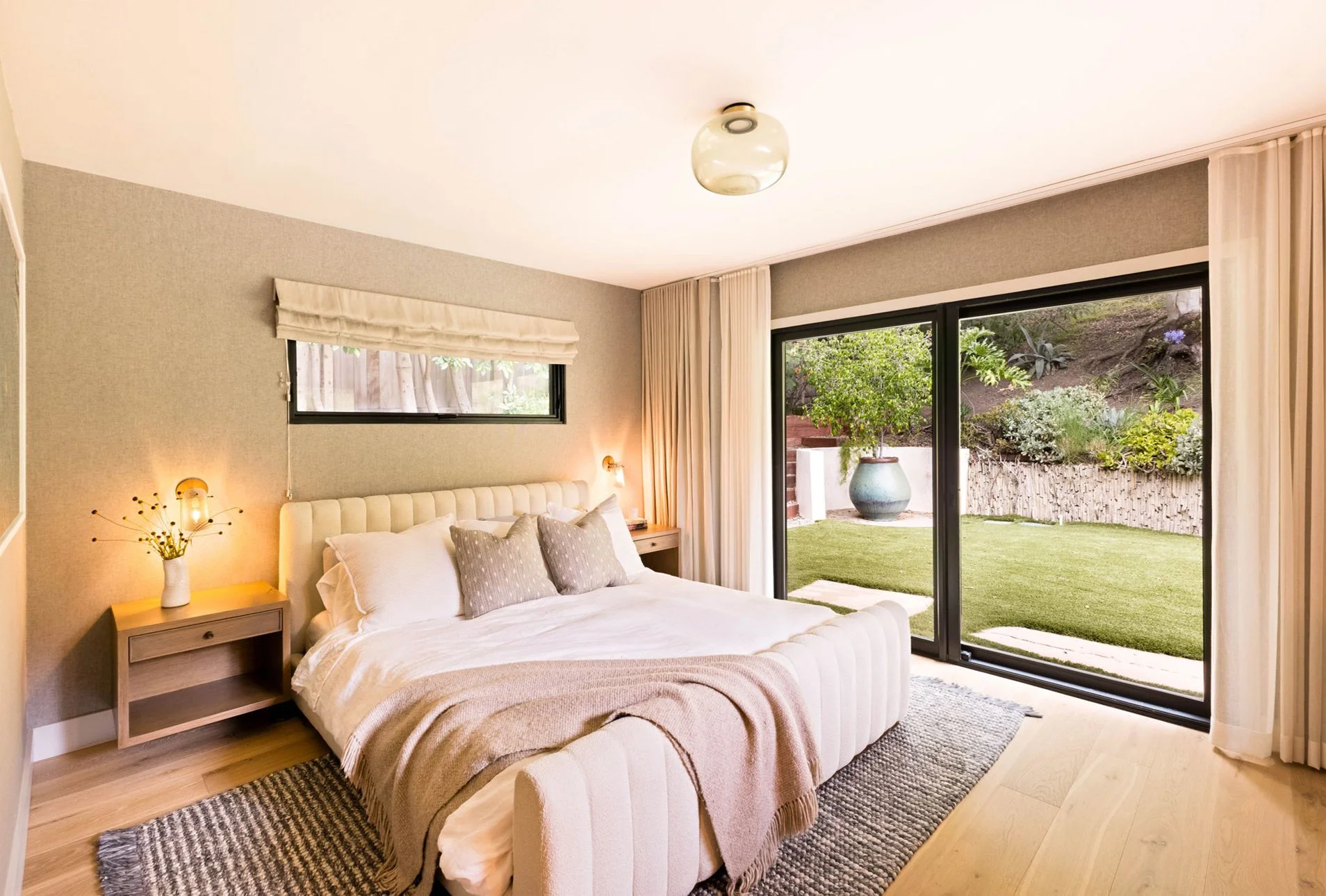 A bright and modern primary bedroom with grey wallpaper, a white boucle channeled bed sits ontop of a chunky area rug. Floating oak nightstands and brass sonces flank the bed.