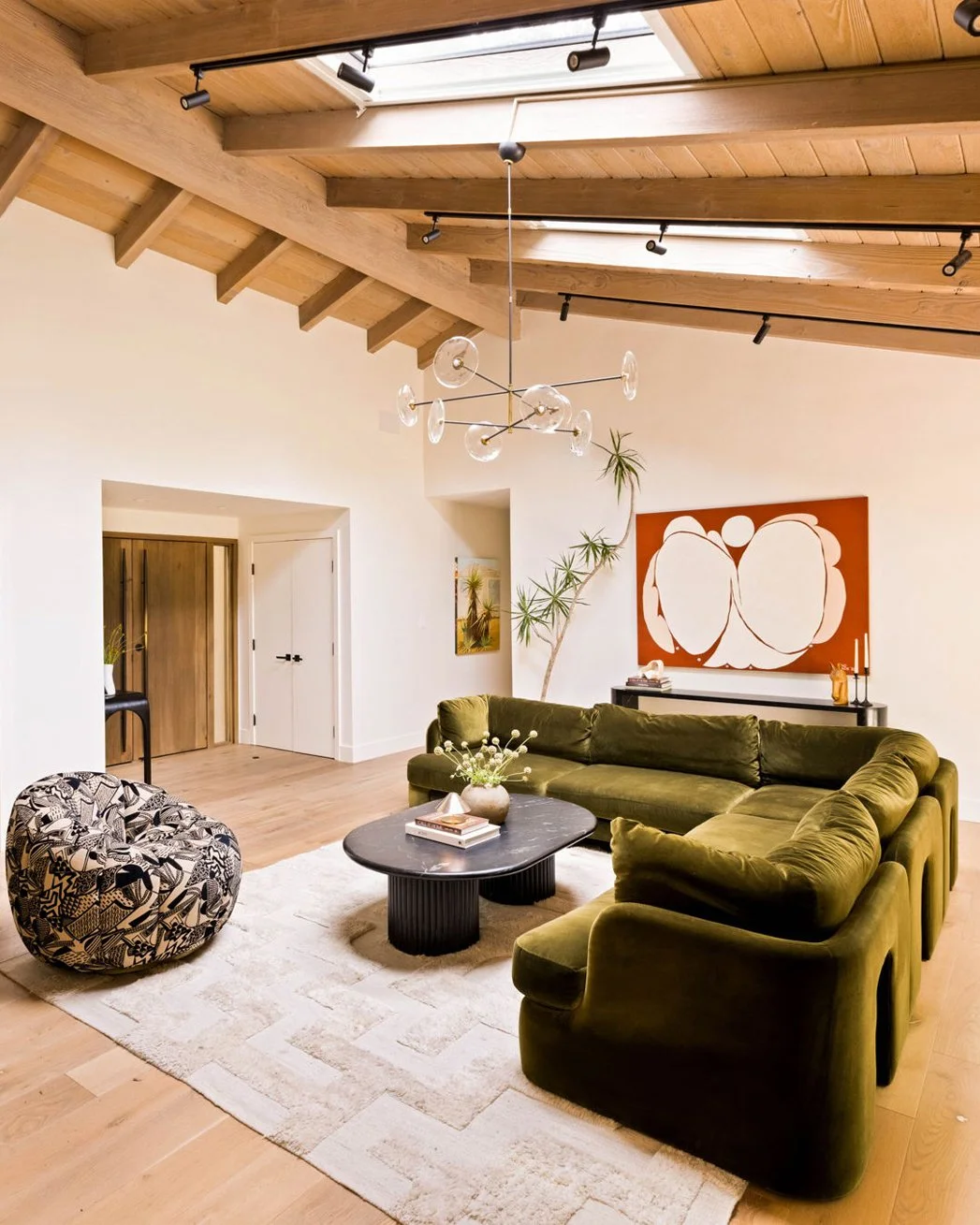 A living room in a mid-century modern home with vaulted wood ceilings and white walls.  An olive-green sectional sofa in anchored by a cream rug with a black marble coffee table.  A red abstract painting and console table are in the background.