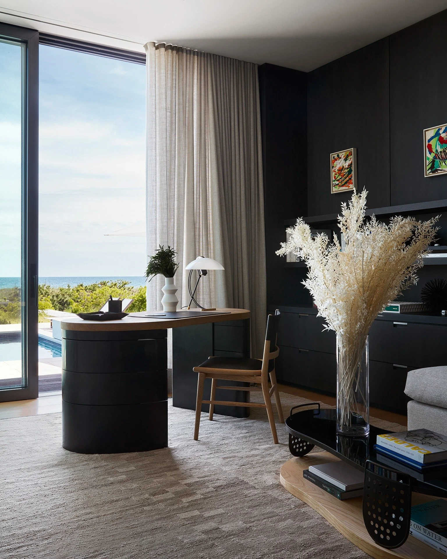 Modern living room with large glass door showing ocean view, black shelving unit with books and decorations, tall white flowers in a vase, and a wooden desk with a lamp and small plant.