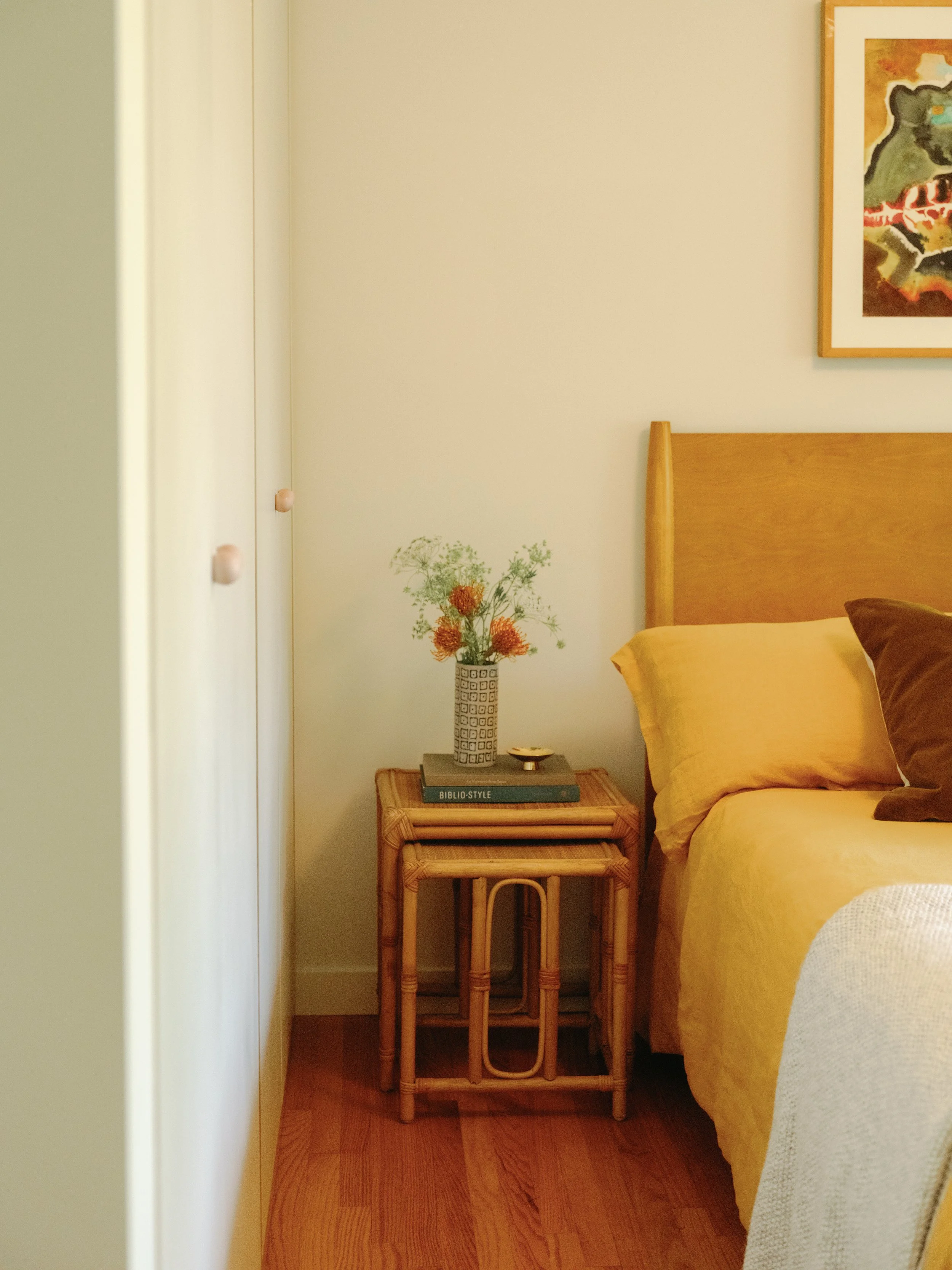 A cozy bedroom corner with a wooden bed frame, yellow bedding, a bedside table with a flower vase, books, and a framed artwork on the wall.