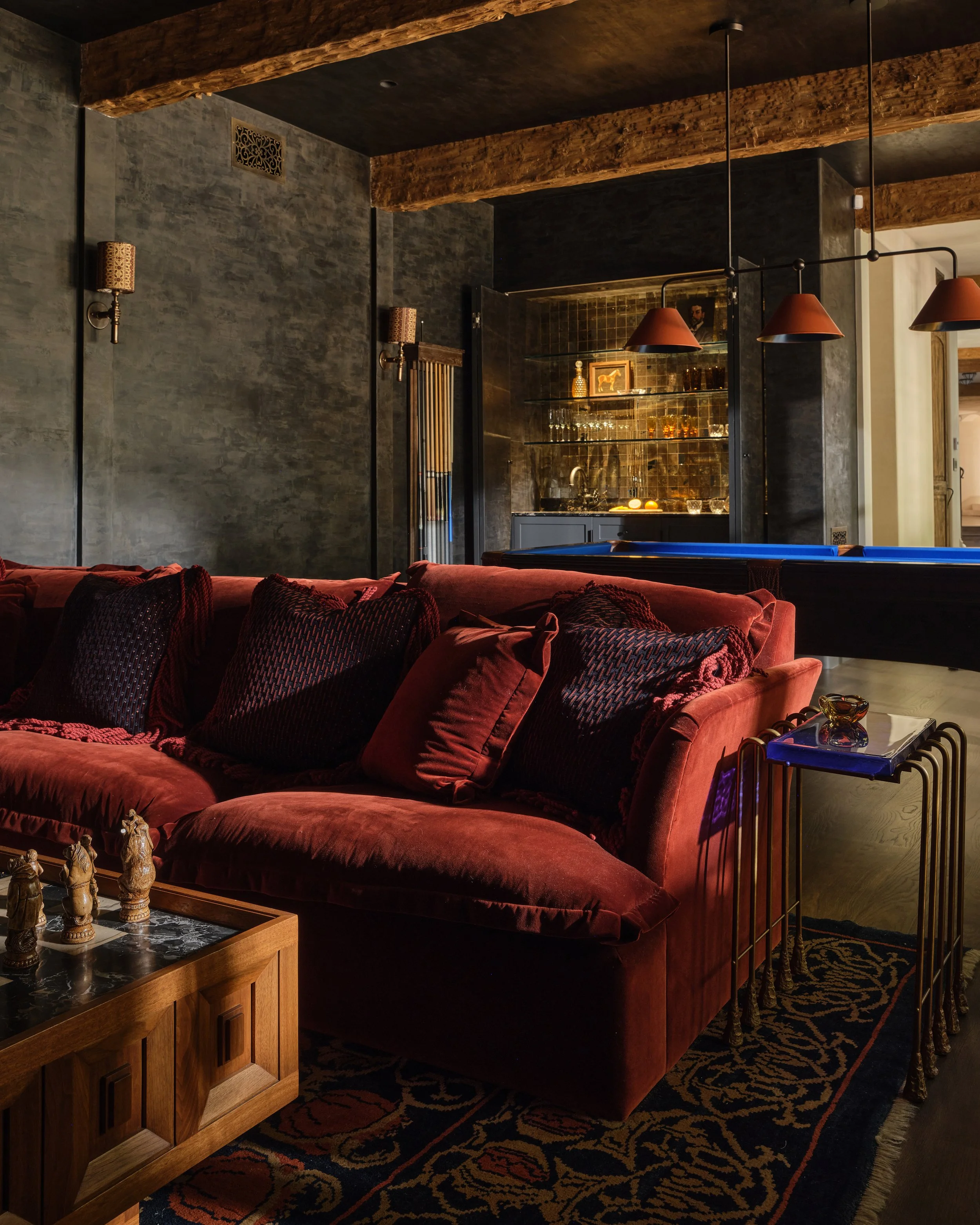 A cozy living room with a red velvet sofa, dark walls, and a bar area in the background with glass shelves and bottles, decorated with warm lighting.