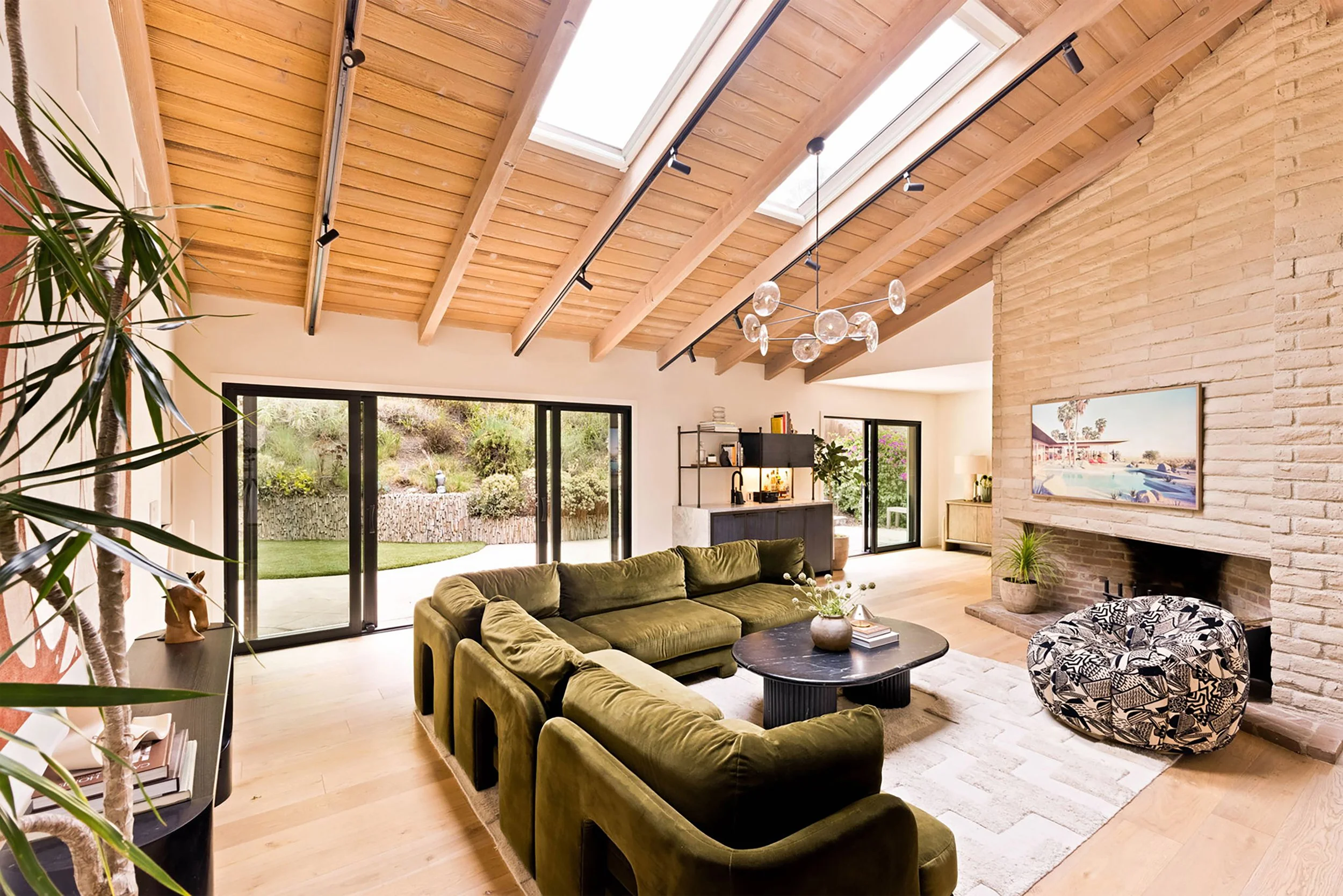 A living room in a mid-century modern home with vaulted wood ceilings. An olive-green sectional sofa in anchored by a cream rug with a black marble coffee table.  A black and white patterned lounge chair sits in front of a brick fireplace.