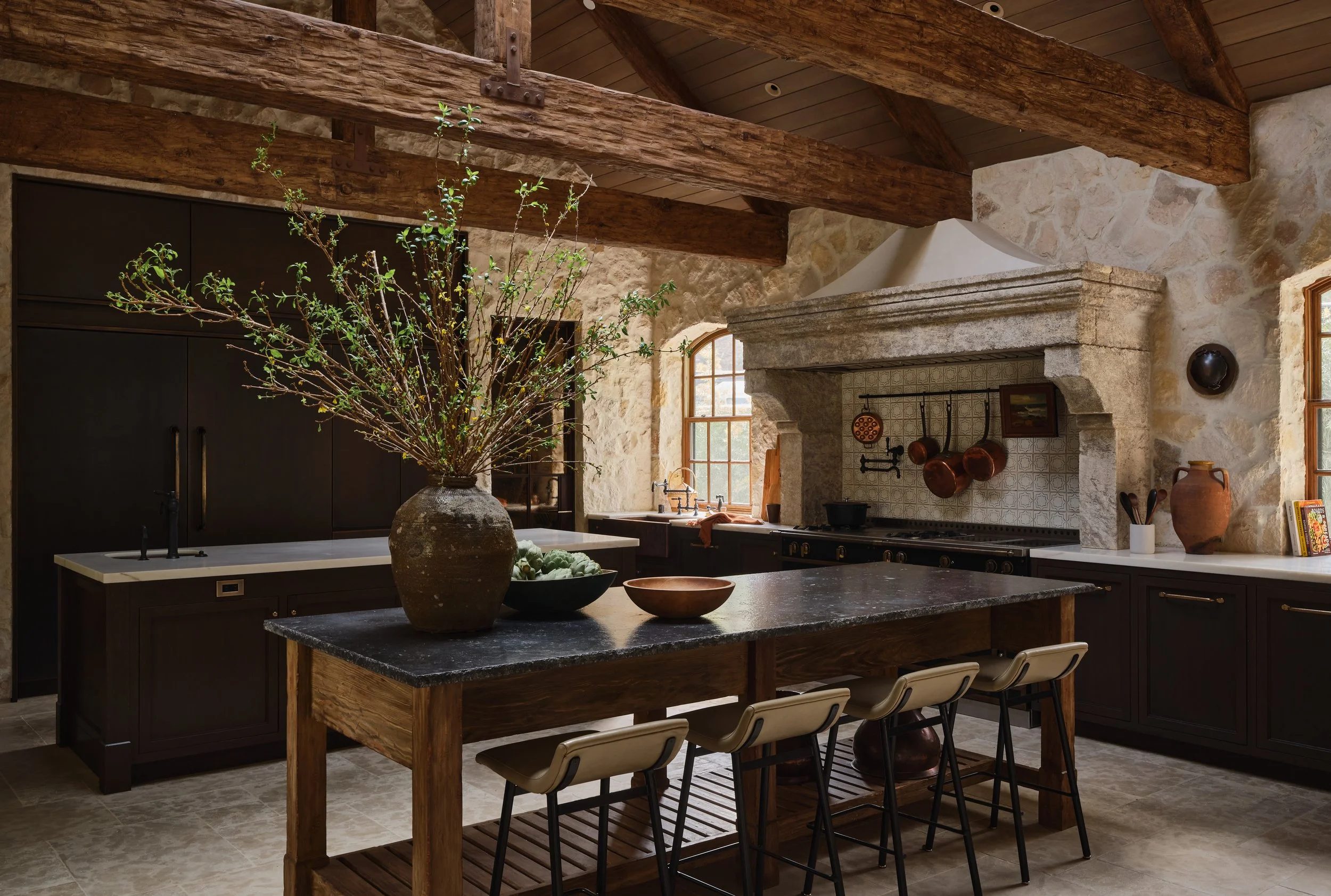 Rustic kitchen with stone walls, wooden beams, black cabinets, a large dark countertop island with a vase of branches, and a stove with copper pots hanging above.