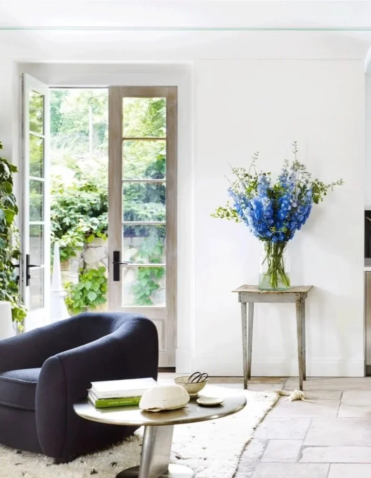 Living room with open glass door, black armchair, white side table with books and decor, and a tall vase with blue flowers on a wooden stand.