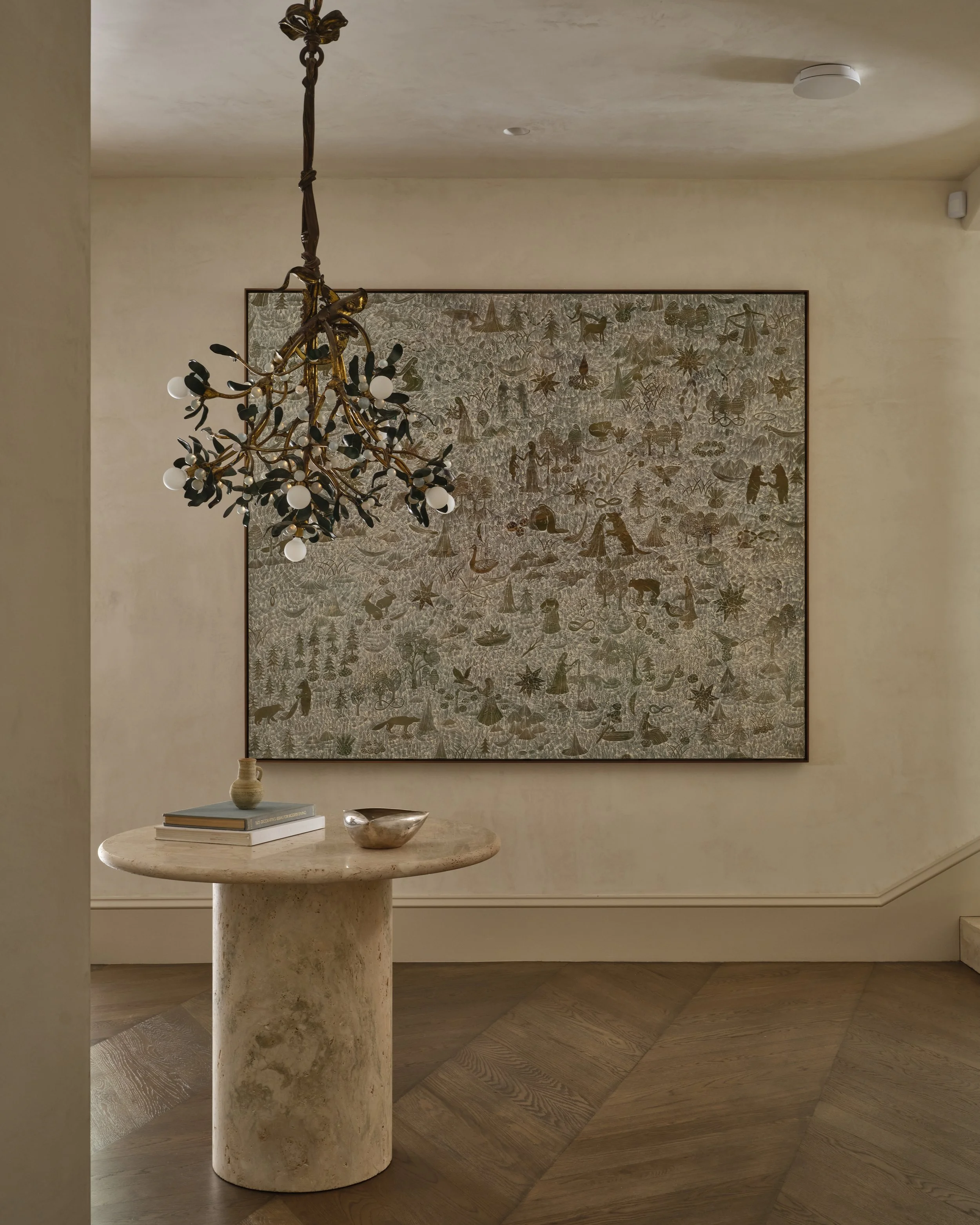 A decorative wall hanging with detailed illustrations, a small marble table with books and a decorative bowl, a gold-colored chandelier with dark accents, and a wooden floor.