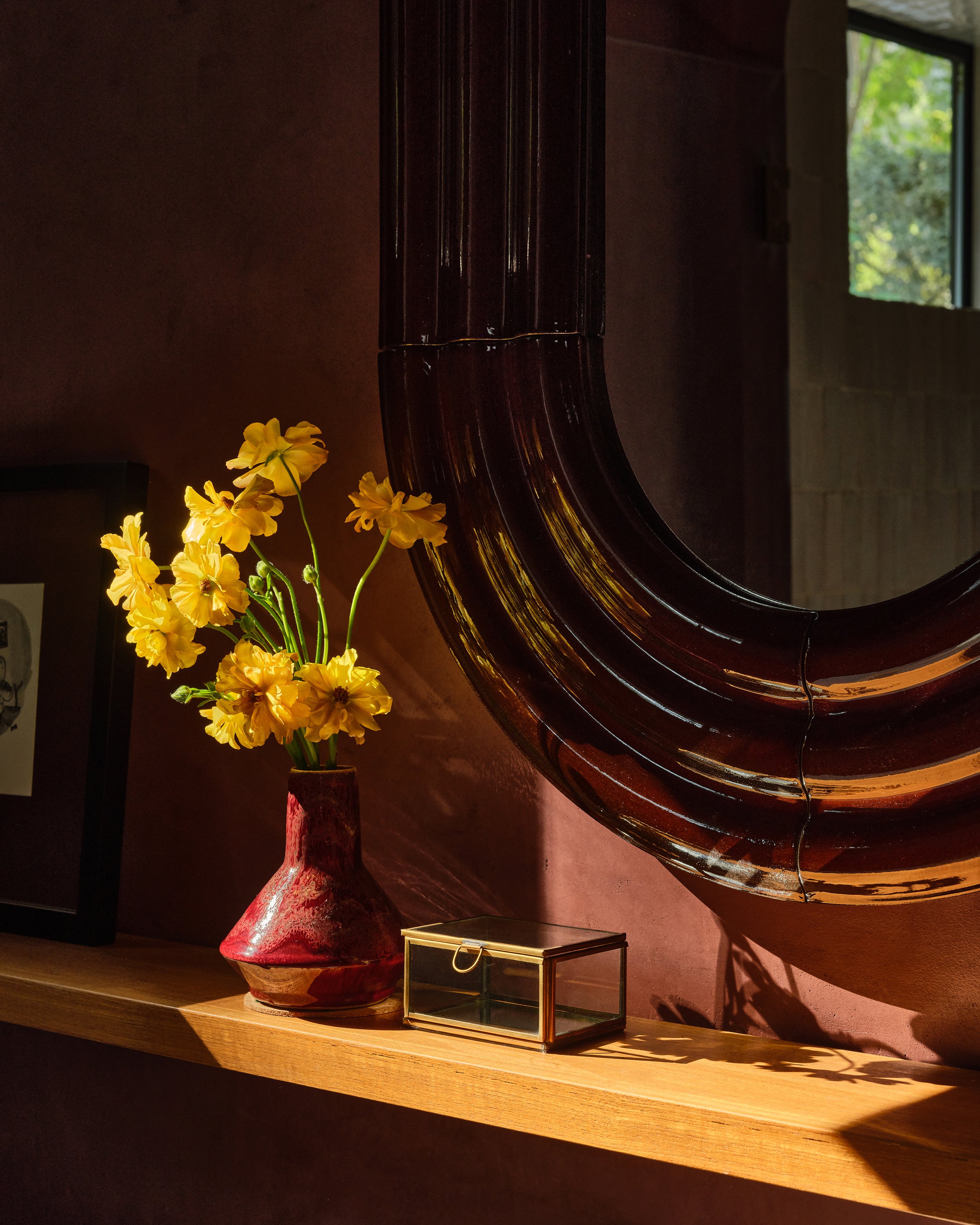 Anthony Jeselnik’s home, interior design by Riccio Vidaurre Design – A bathroom with burgandy plaster walls, an brown ceramic elo mirror by pani jurek, a wooden shelf with a small red ceramic vase with yellow flowers and a small glass box.