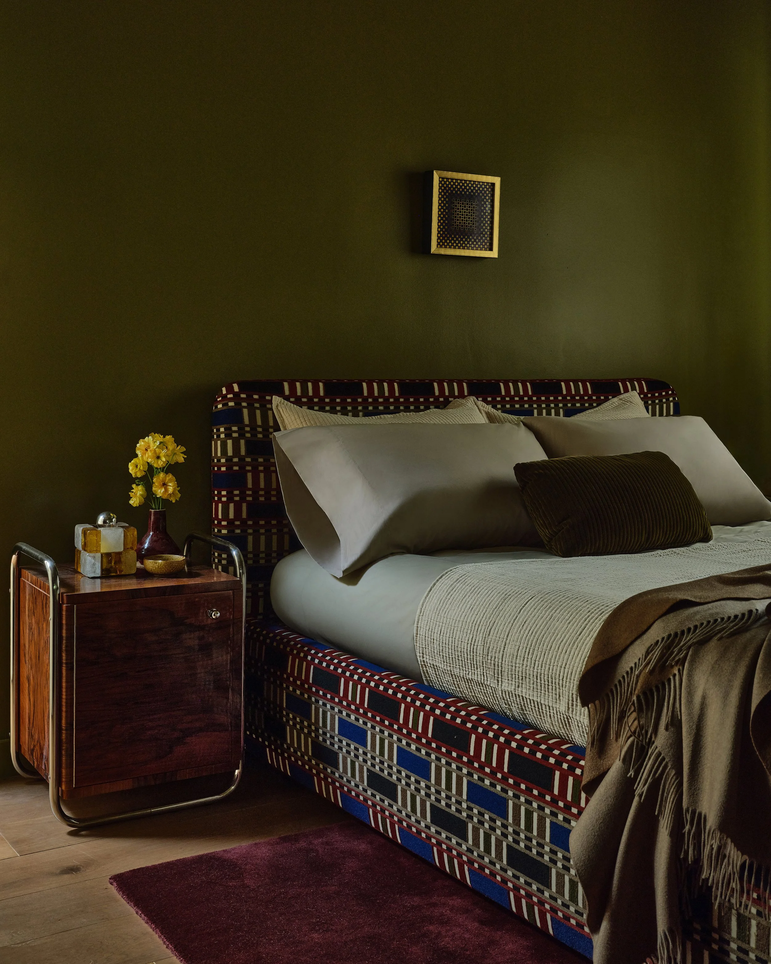 Anthony Jeselnik’s home, interior design by Riccio Vidaurre Design – A guest bedroom with walls painted olive green, a custom bed upholstered in limpop pierre frey fabric, a vintage  walnut and chrome nightstand and a piece of art by victor vasarely.