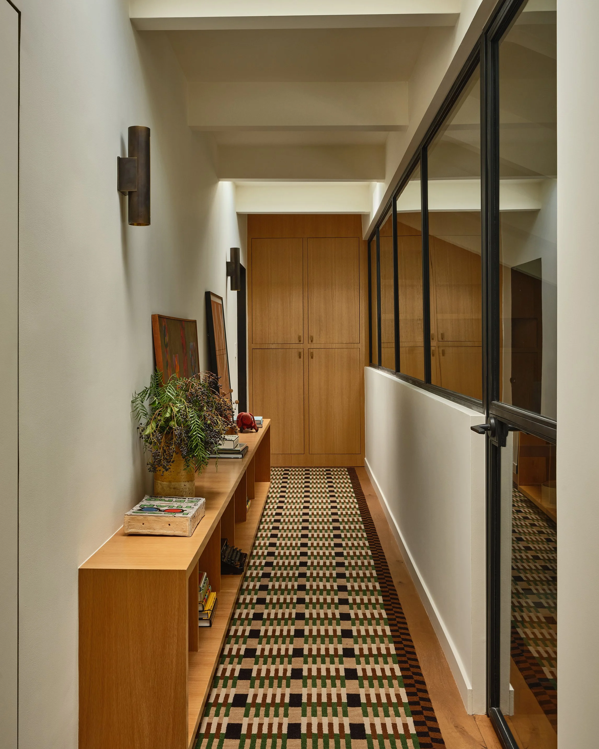 Anthony Jeselnik’s home, interior design by Riccio Vidaurre Design – the hallway with a custom floor runner by erik lindstrom, with a wooden built-in console and closet, sconces above and a wall of windows.