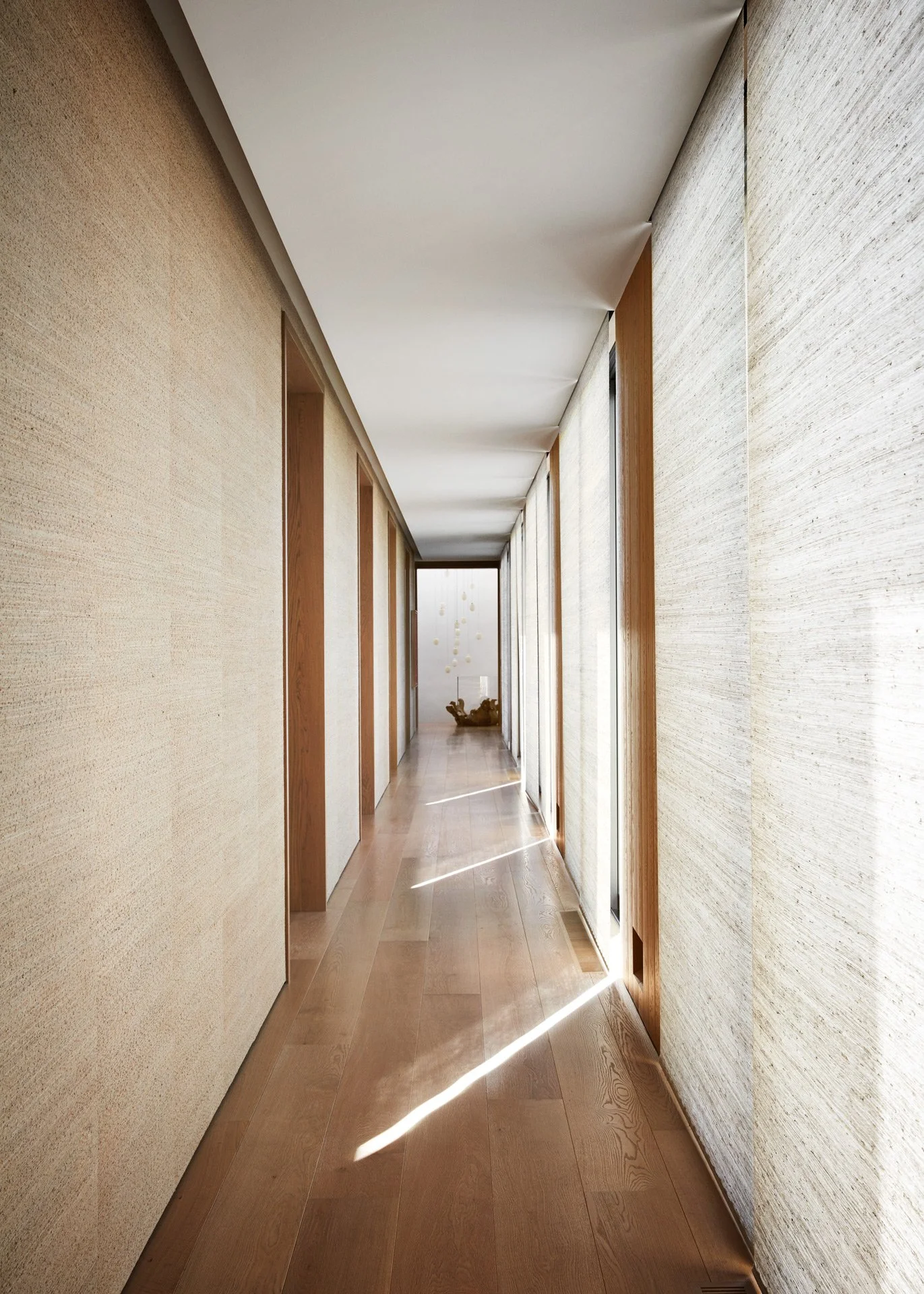 A long, narrow hallway with wooden flooring and textured walls on either side, lit by sunlight streaming through tall windows with blinds, leading to a door with decorative yellow dots and a sculpture at the end.