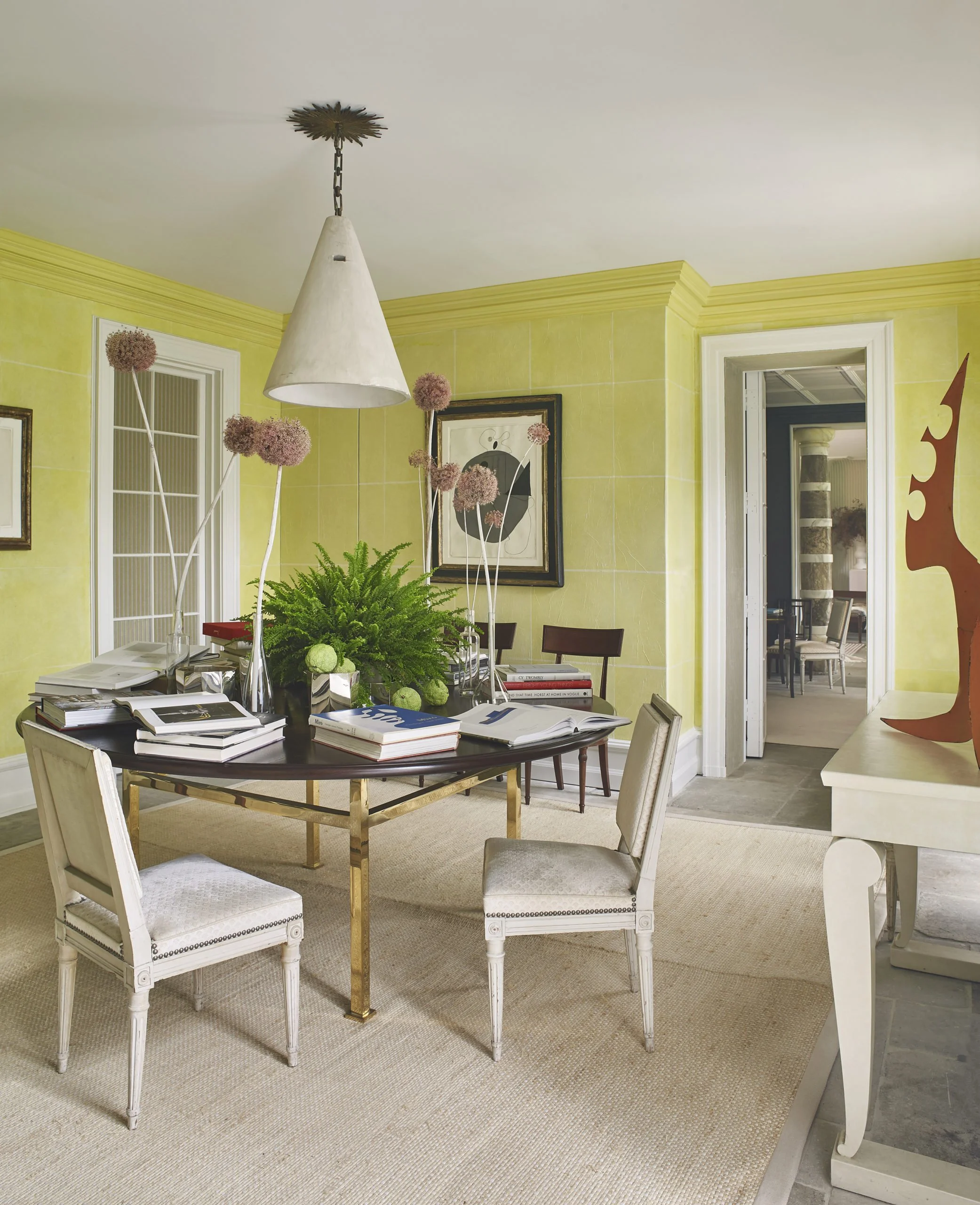 A dining room with yellow walls, a round table with stacks of books, a floral centerpiece, a white pendant lamp, beige chairs, and a doorway leading to another room with a striped column.