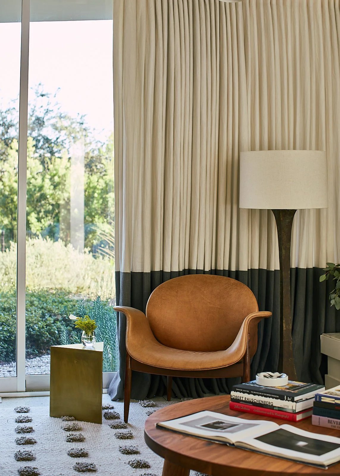 A cozy living room corner with a tan leather armchair, a side table with a small plant, floor-to-ceiling curtains, a window showing greenery outside, a tall floor lamp, a round wooden table with open books and magazines.