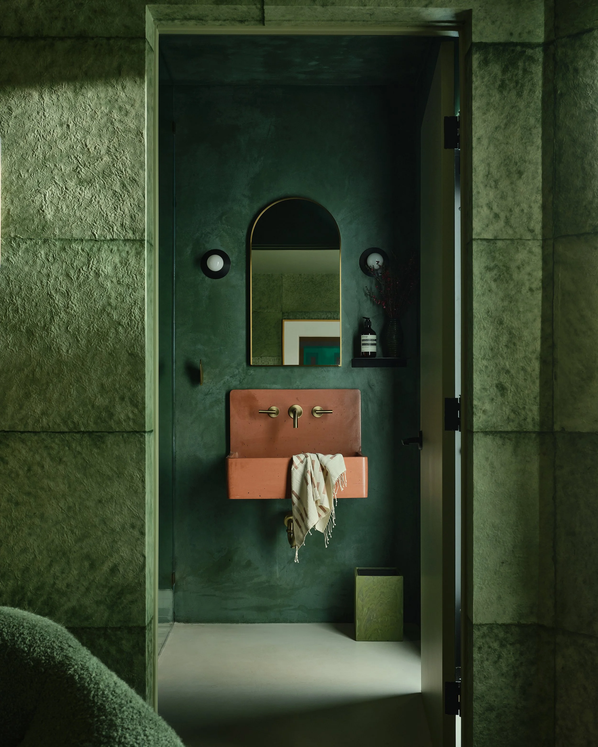 Comedian Anthony Jeselnik's home by Riccio Vidaurre Design.  A monochromatic teal green bathroom with green plaster walls and a orange concrete sink by kast.  An arched mirror hangs over the sink flanked by two black sconces.