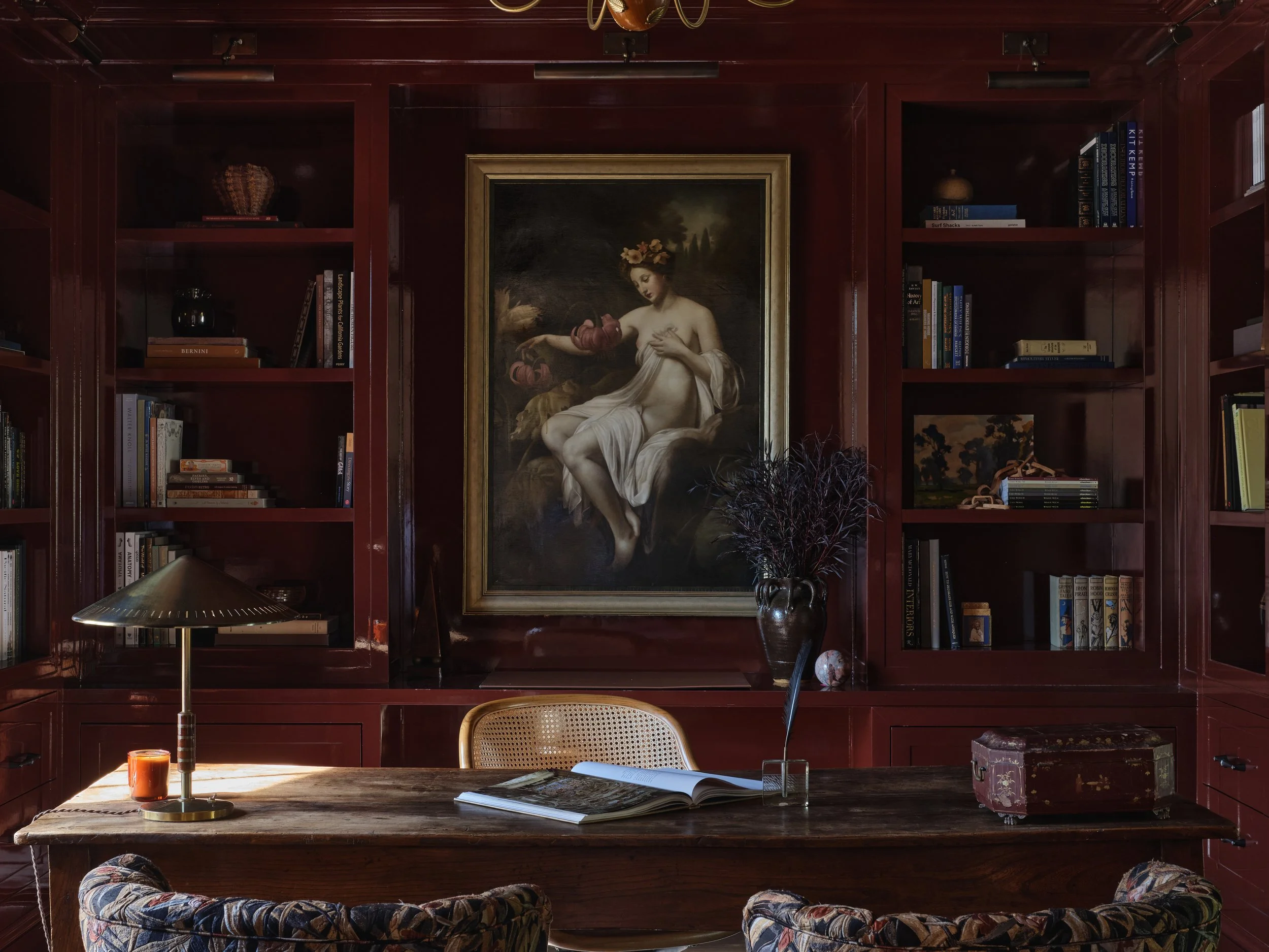 A room with dark red bookshelves, a large classical painting of a woman with flowing white dress, and a wooden desk with open book, table lamp, and decorative objects.