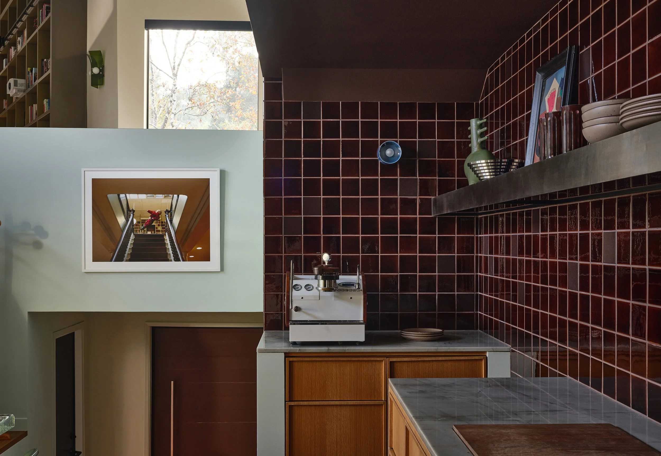 Comedian Anthony Jeselnik’s home by Riccio Vidaurre Design – A kitchen with oak cabinets and brown square ceramic tiles backsplash with a stainless-steel shelf styled with objects.  A photograph hangs above an entryway.