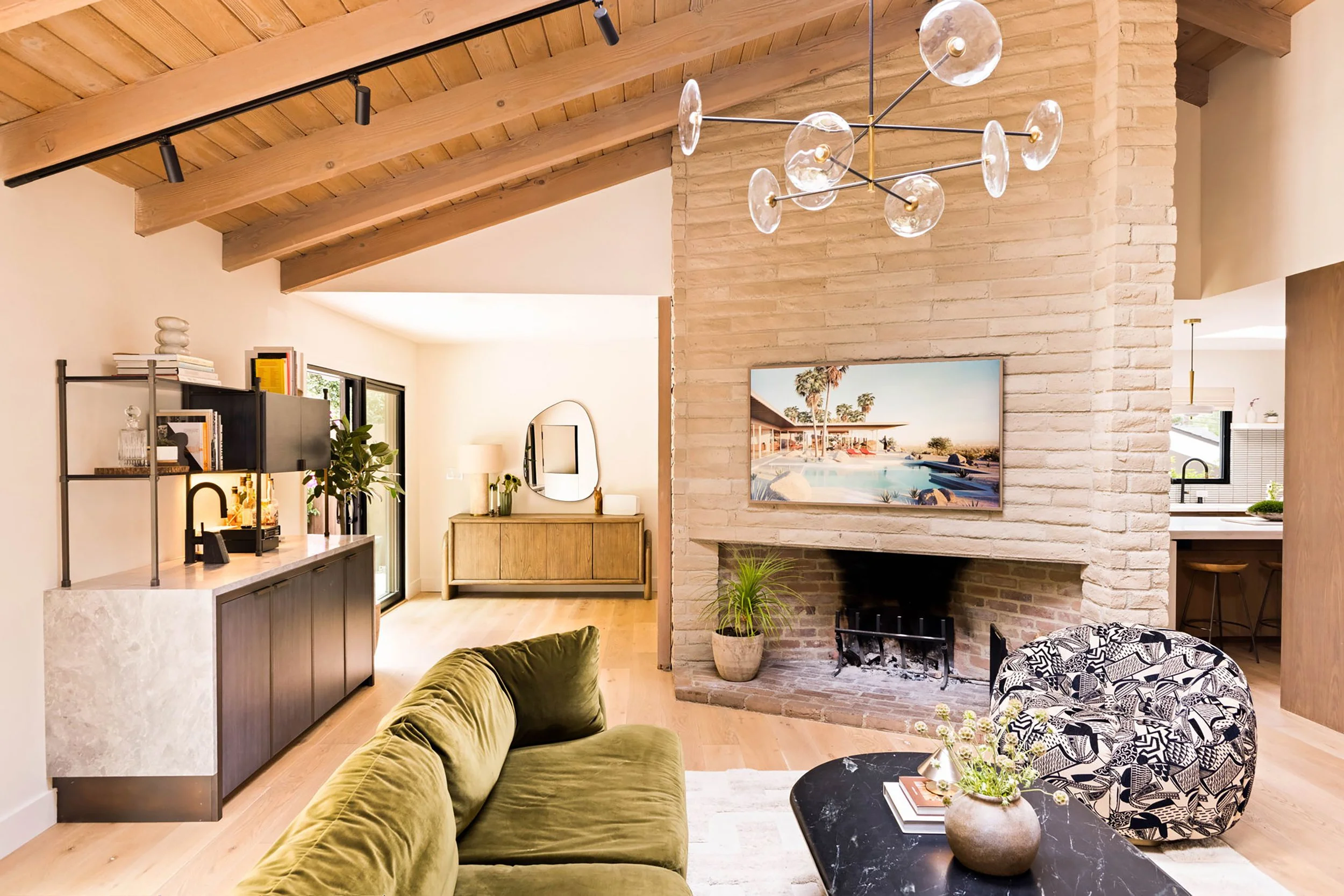 A cream brick fireplace in a mid-century modern home with vaulted wood ceilings. An olive-green sofa, black marble coffee table and black and white patterned lounge chair sit under a modern glass chandelier.