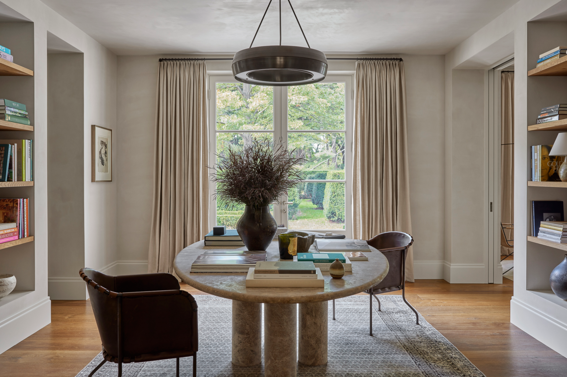 A cozy dining room with a round table, two brown chairs, a large vase with dried branches, and a view of a lush green garden through a window with beige curtains.