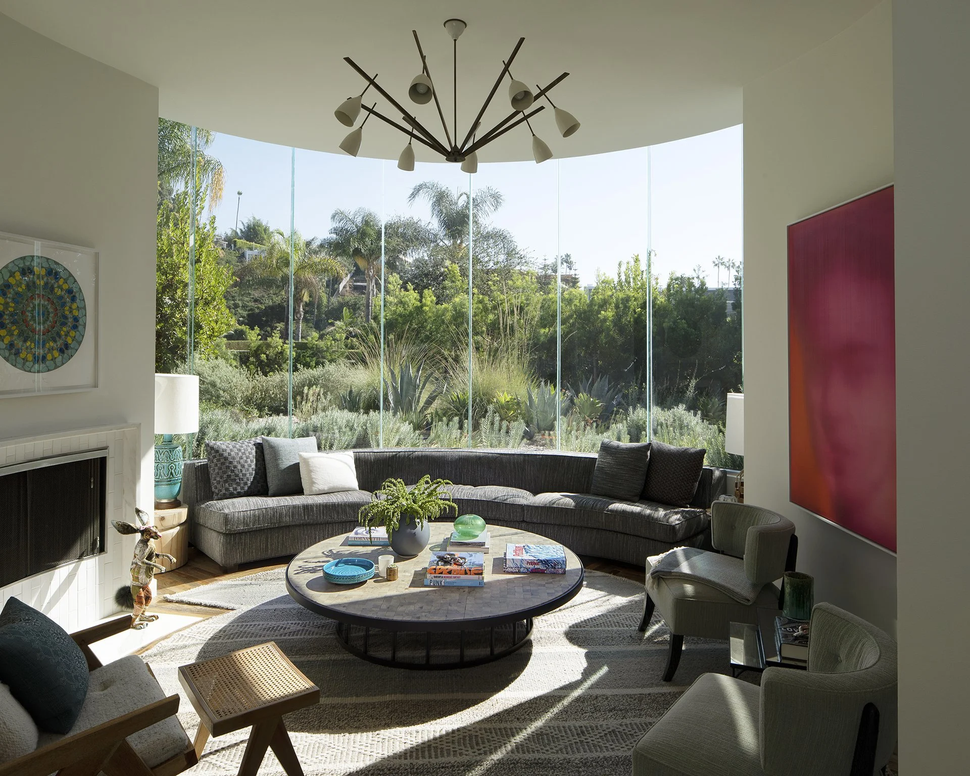 Bright living room with large curved window overlooking a lush garden, modern chandelier, gray sectional sofa with cushions, round coffee table with books and plant, armchairs, colorful artwork on walls, and side tables with lamps.