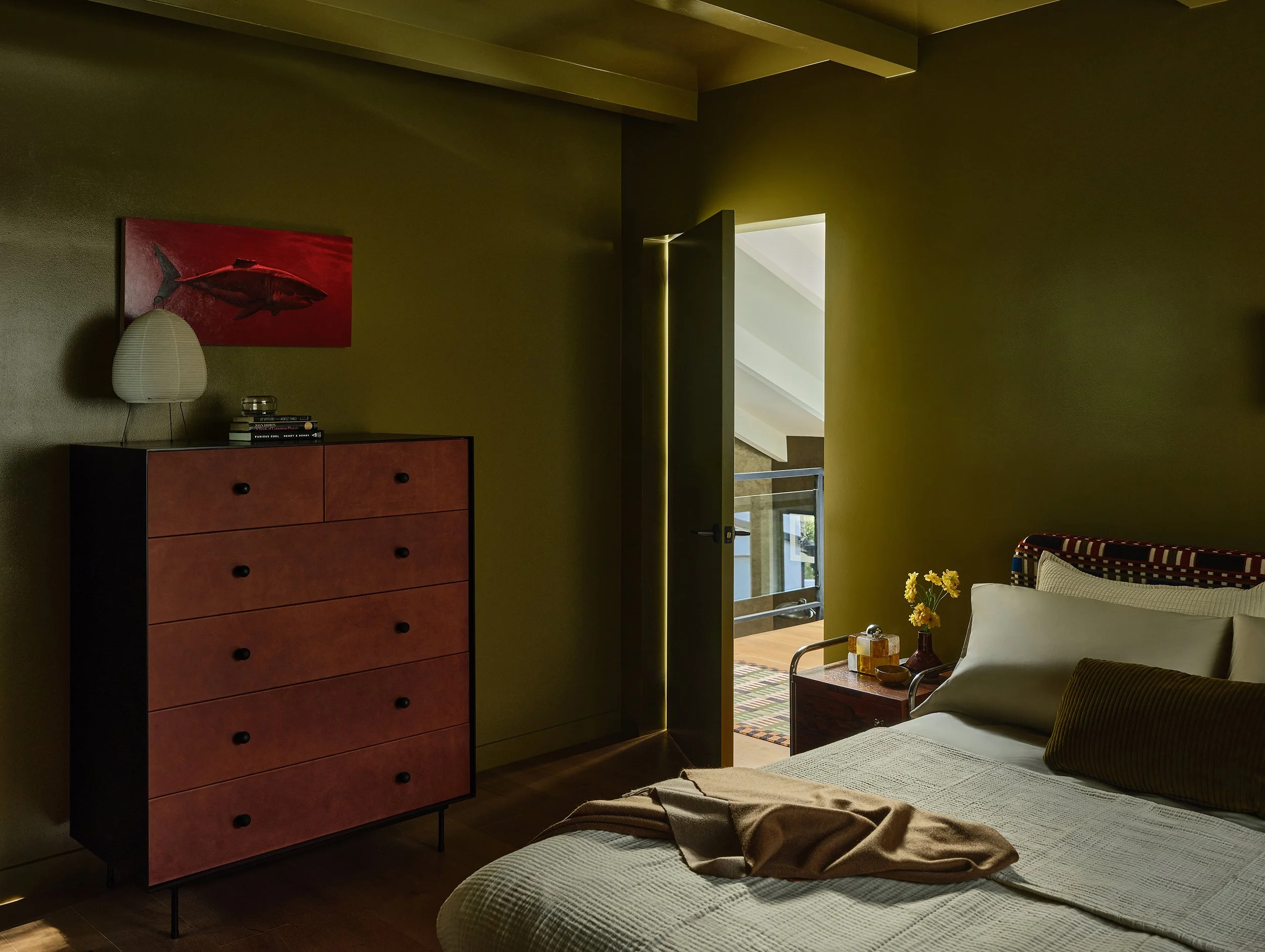 Anthony Jeselnik’s home, interior design by Riccio Vidaurre Design – A bedroom with olive green walls, a black metal and burgandy leather dresser by croft house with a red painting of a shark above and a paper lamp.