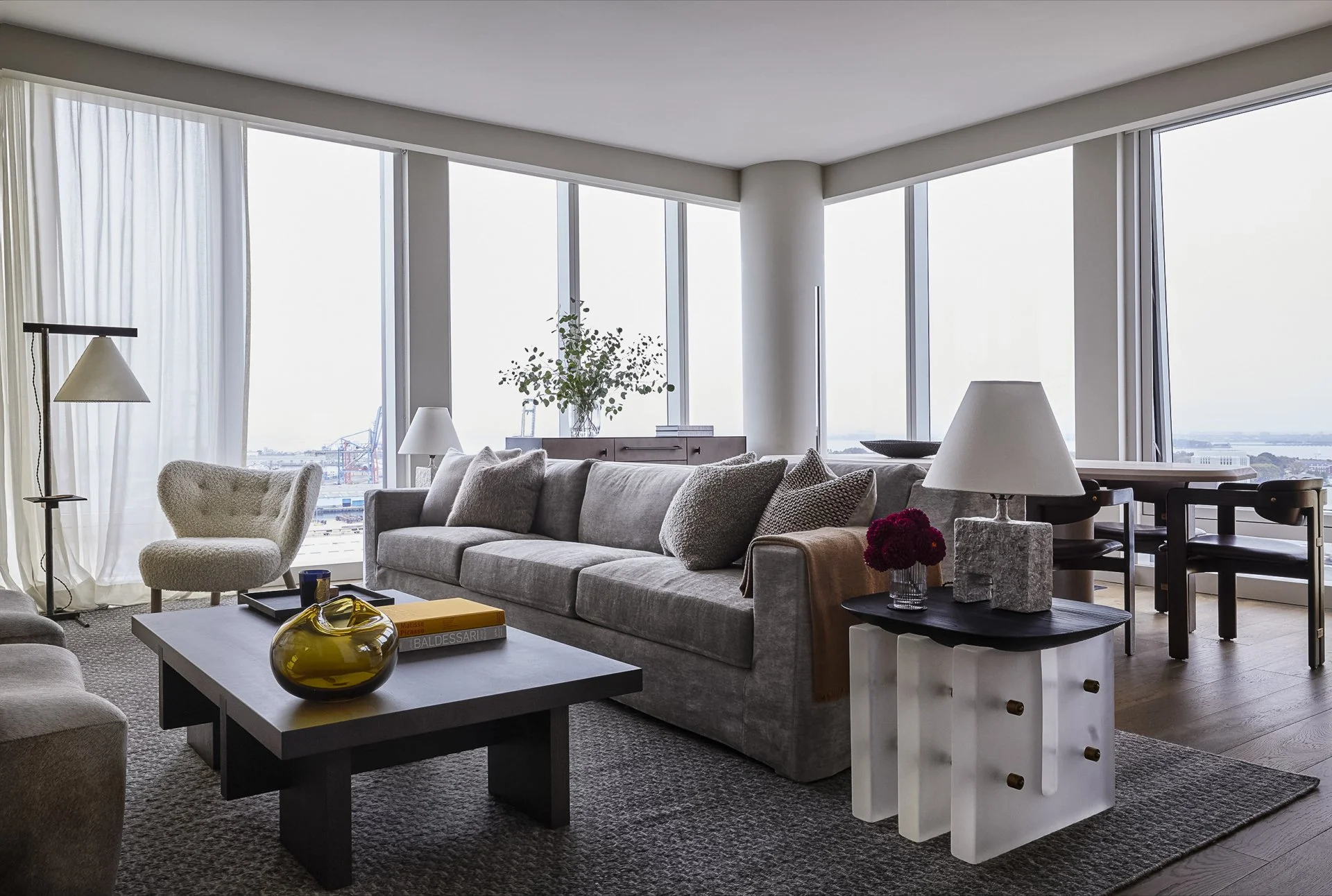 Modern living room with large windows overlooking a cityscape, featuring a gray sofa, beige armchair, coffee table with decorative items, side table with a lamp and flowers, and a dining area in the background.