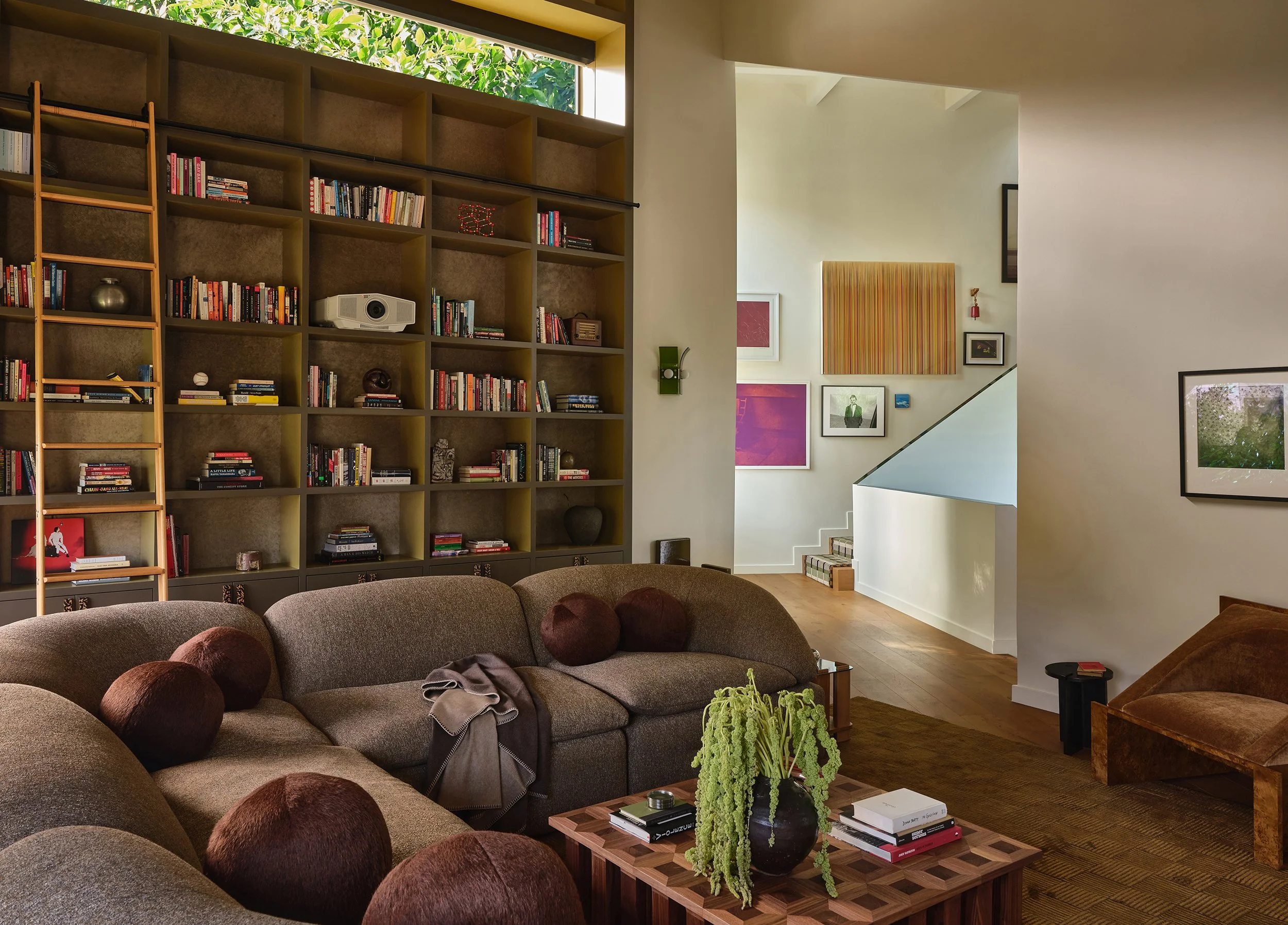 Comedian Anthony Jeselnik’s living room, interior design by Riccio Vidaurre Design – A large brown sectional butter sofa by faye toogood and a tessellated wood coffee table sit in front of a olive green bookshelf with a rolling library ladder.