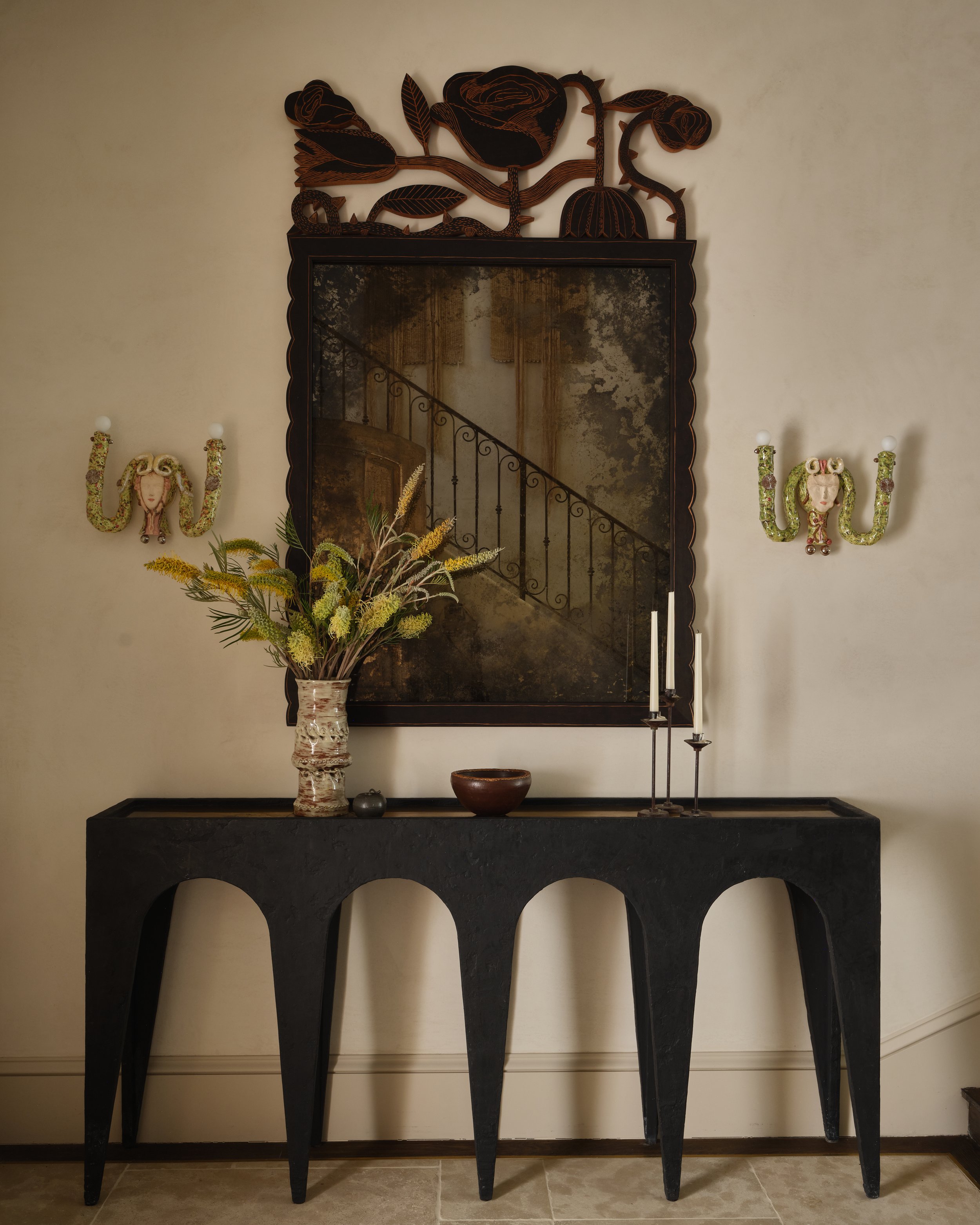 A black console table with arched legs holding a vase of dried flowers, a small bowl, and two tall candlesticks. Above the table is a large mirror with a dark wooden carved floral frame, reflecting a staircase and wall. Flanking the mirror are two decorative wall sconces shaped like women's faces with floral designs and curving arms.