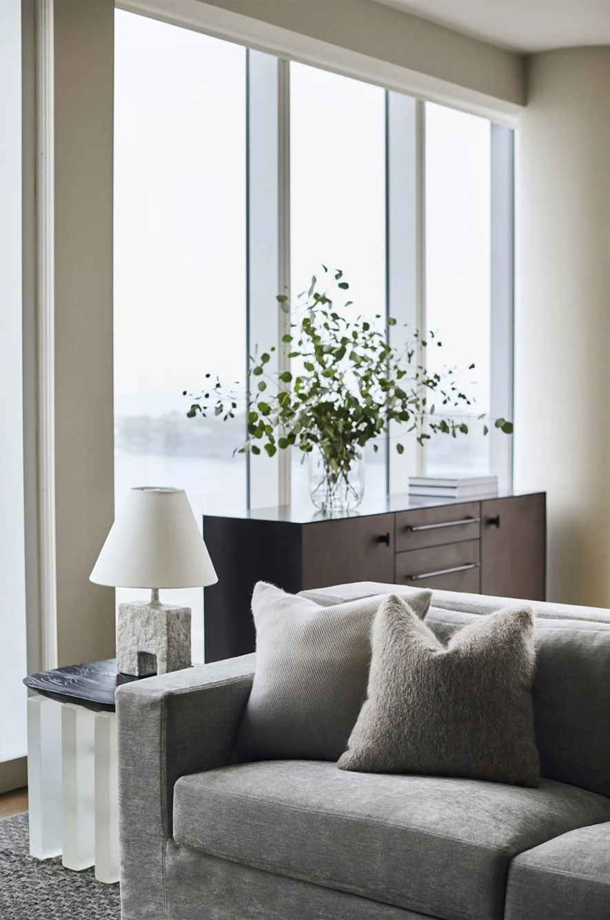 Contemporary living room with large windows, a gray sofa with textured pillows, a dark wooden sideboard with a glass vase filled with green leafy branches, a table lamp on a small table, and stacked books on the sideboard.